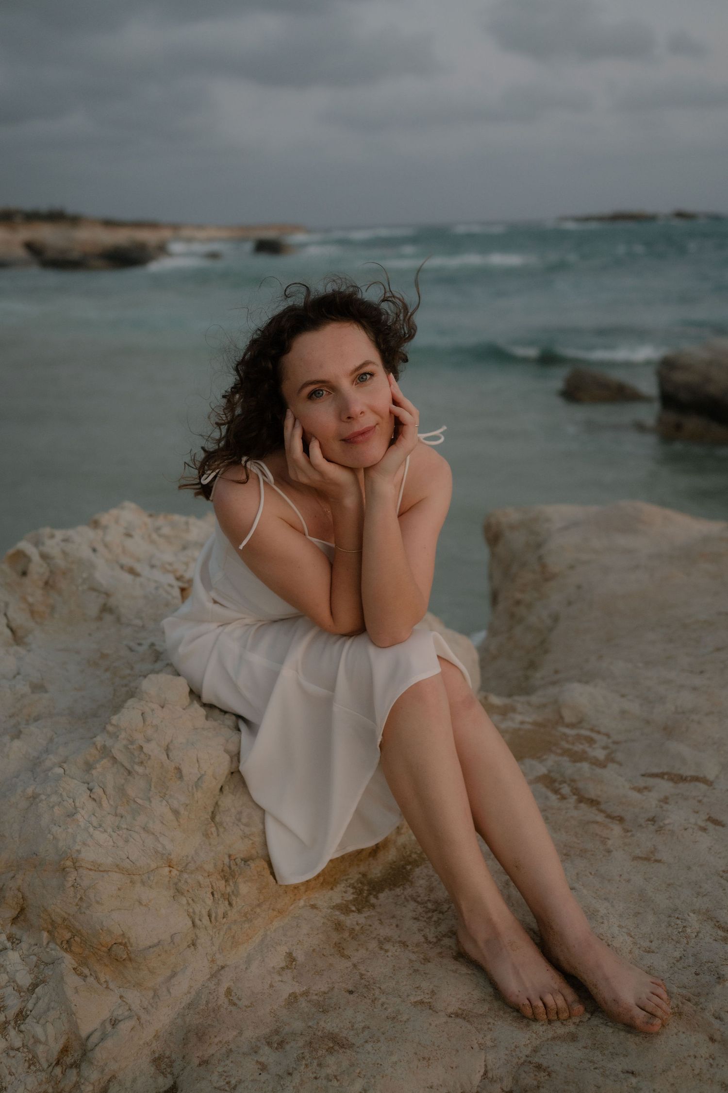 A woman in a white dress sits barefoot on rocky shore, hands framing her face, with turquoise sea behind her.