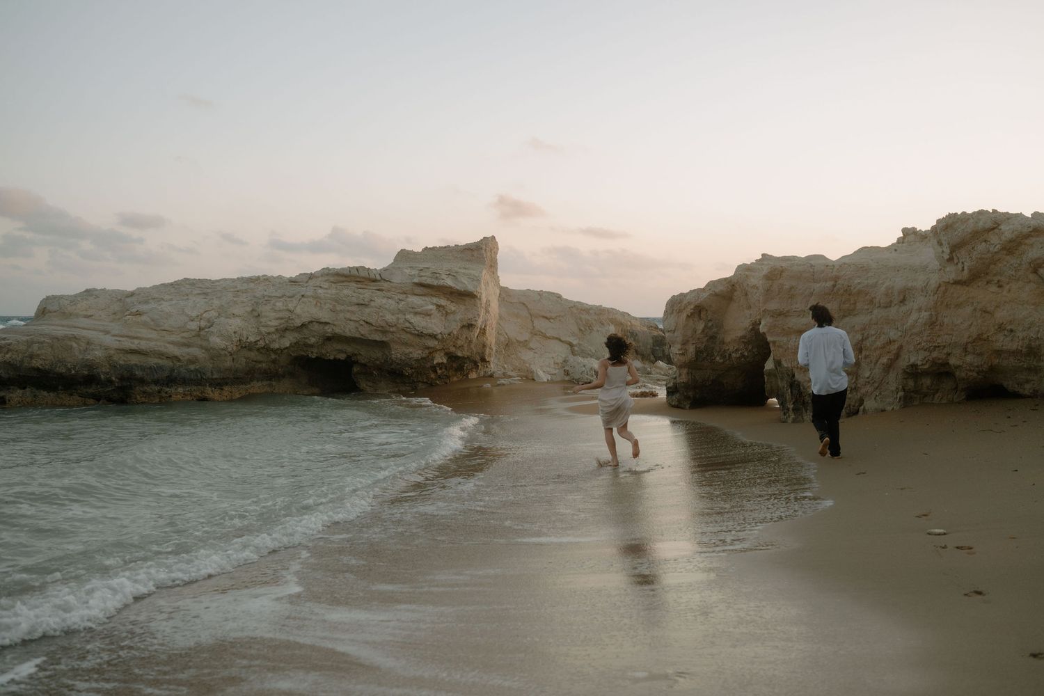 Two people walking on a sandy beach near rocky cliffs at sunset with gentle ocean waves.