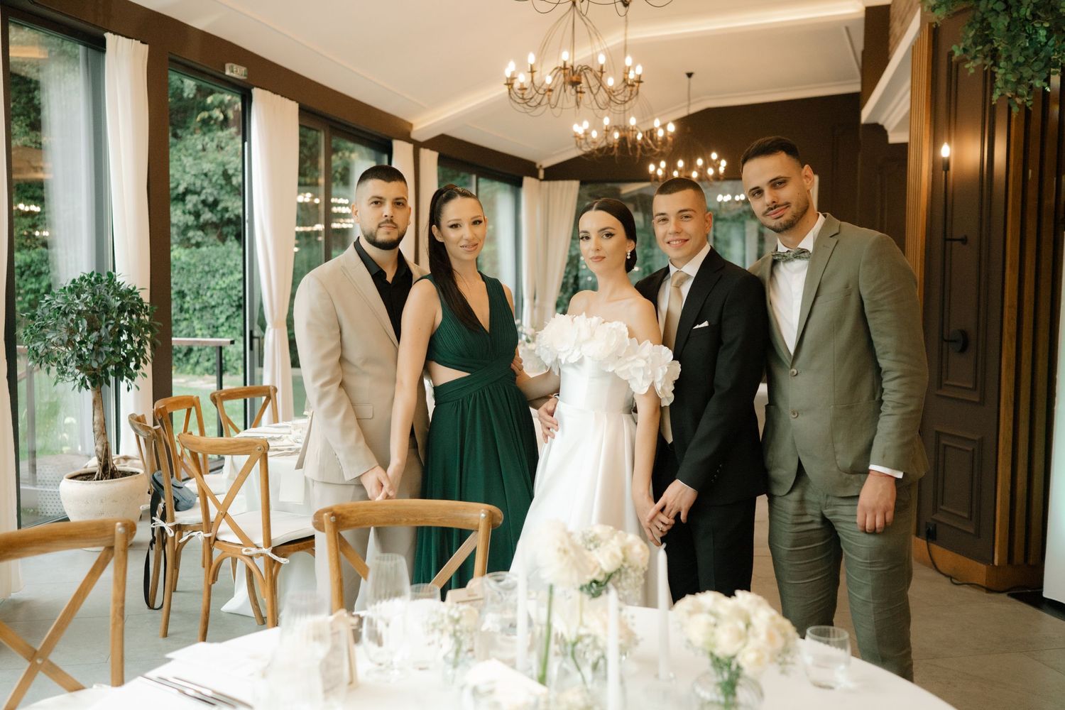 Wedding party gathers around an elegant white wedding cake at an indoor reception venue with chandeliers and greenery.