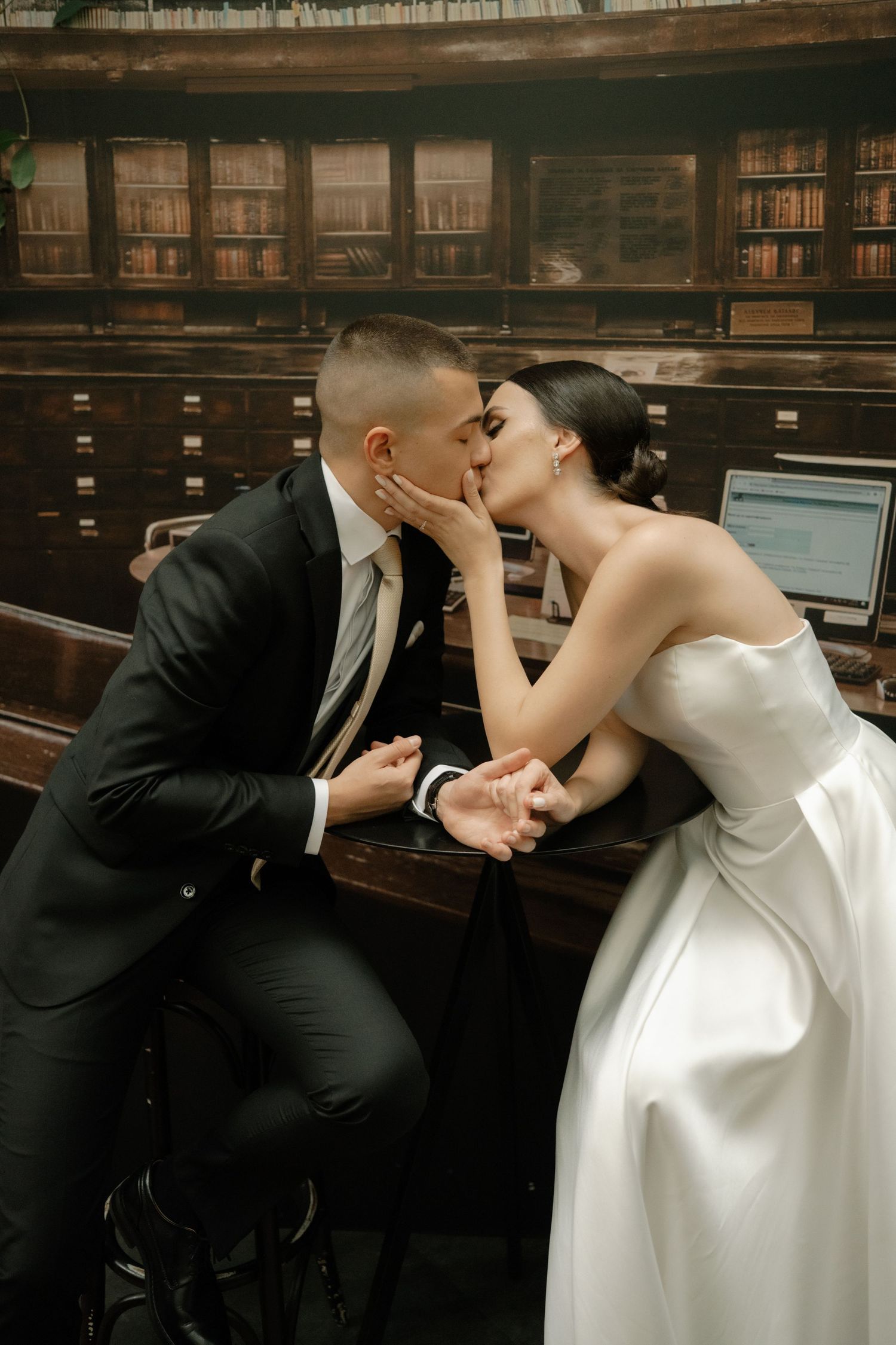 A romantic wedding couple shares an intimate kiss while sitting on a vintage wooden bench in a library setting.