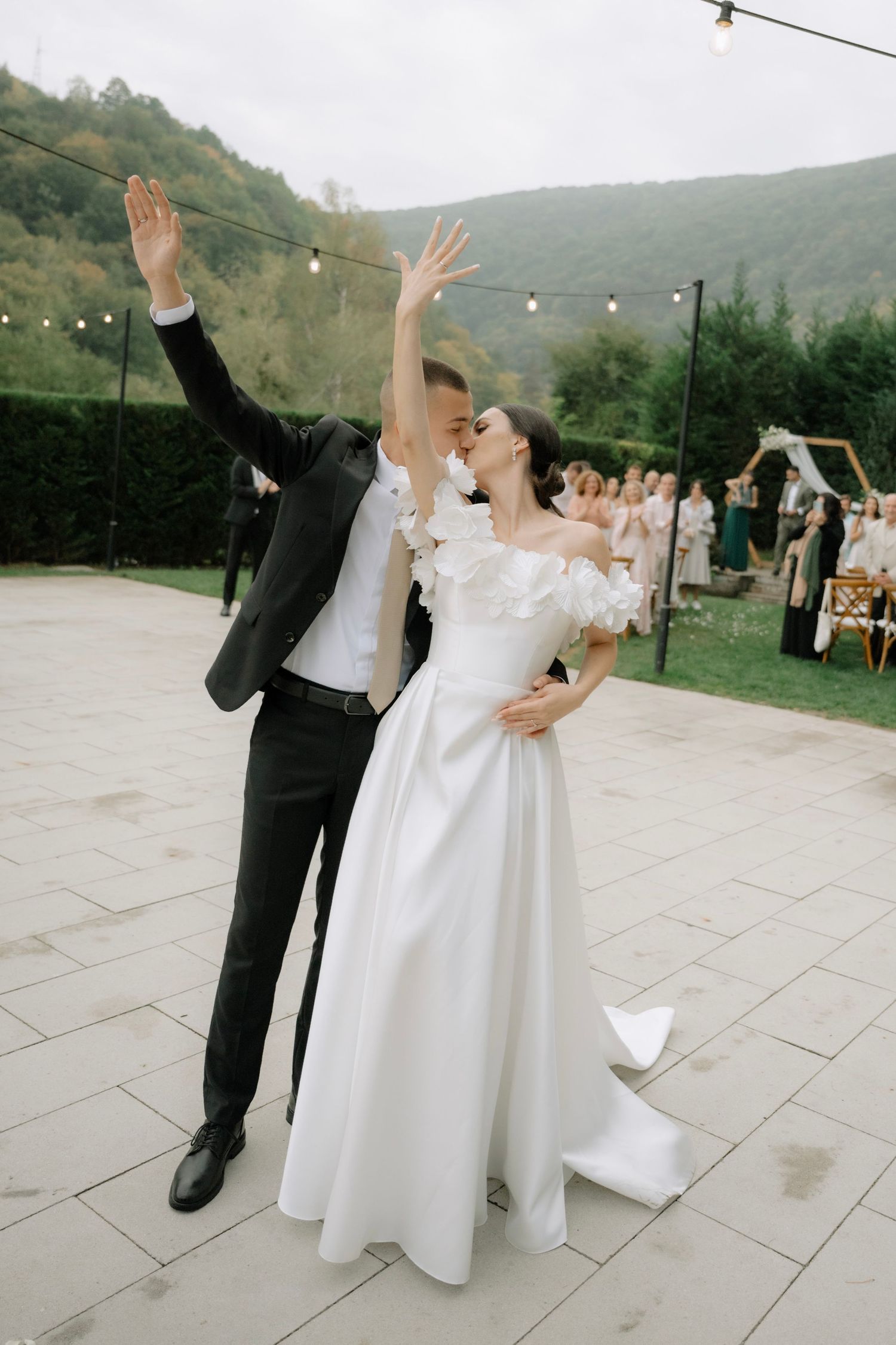 Joyful couple celebrates at outdoor wedding reception on stone patio with mountain backdrop and string lights overhead.