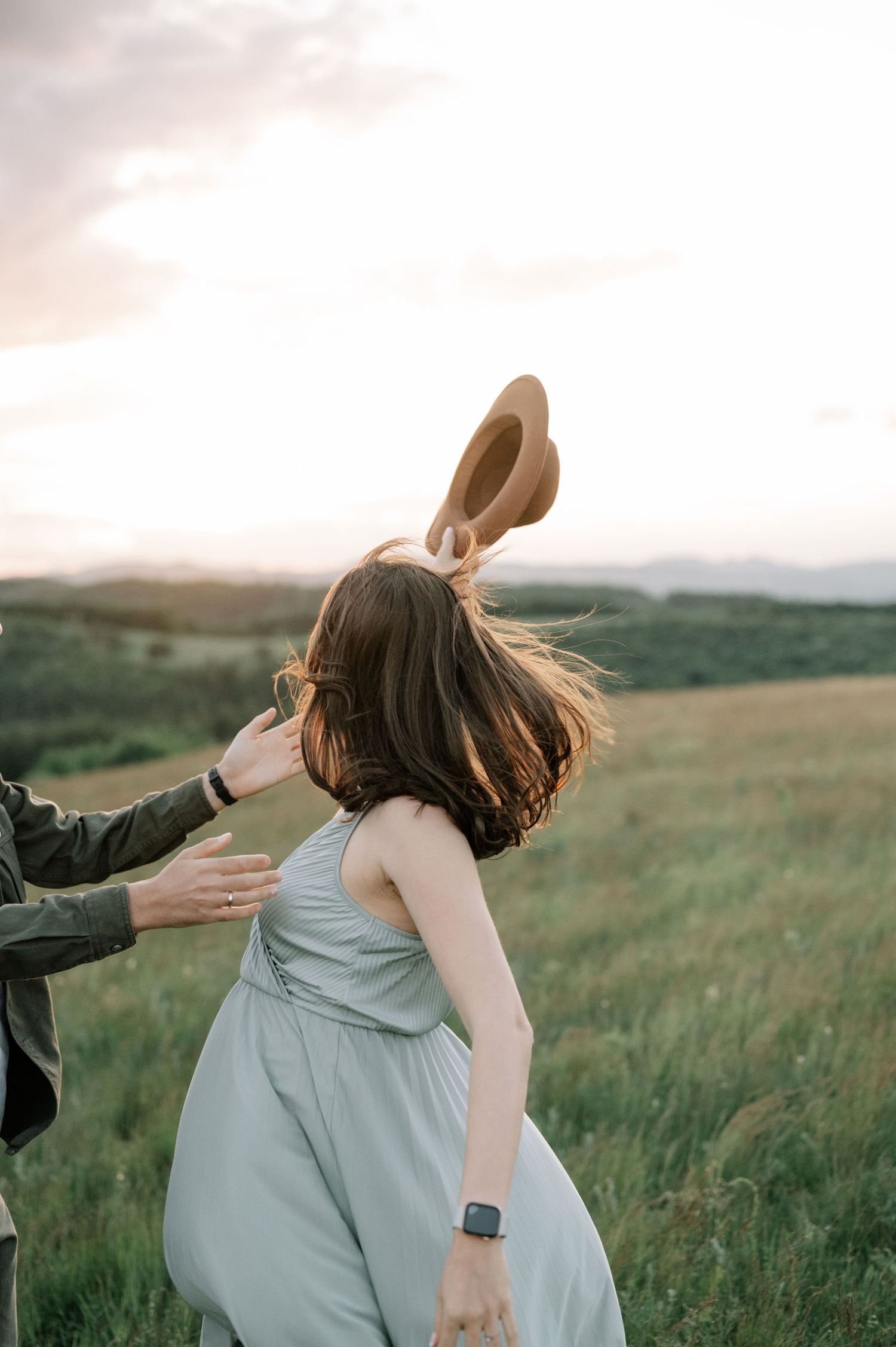 Couple dancing in a green meadow at sunset, woman in light blue dress holding a hat in the wind.