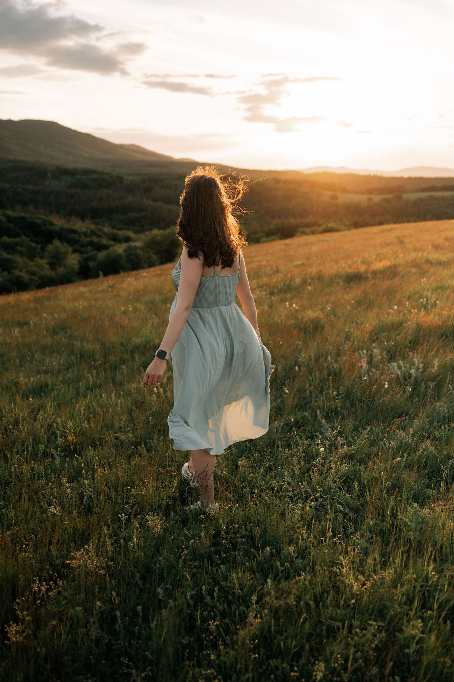 Woman in light blue dress walking through golden meadow at sunset with mountains in background.