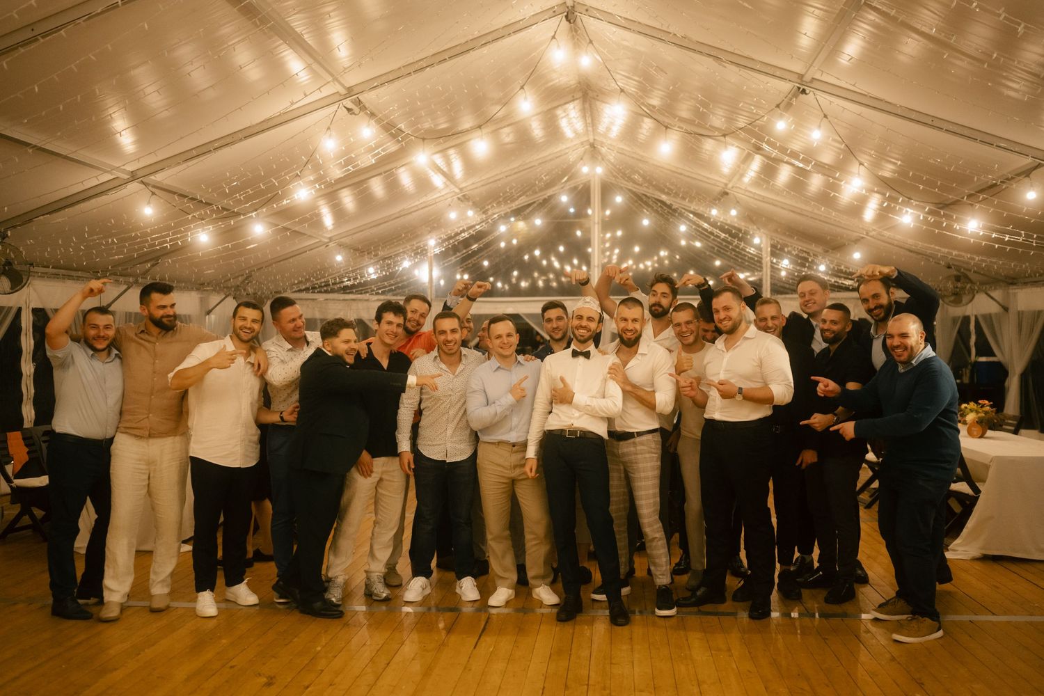 Large group photo of wedding guests gathered under string lights in a rustic reception venue.