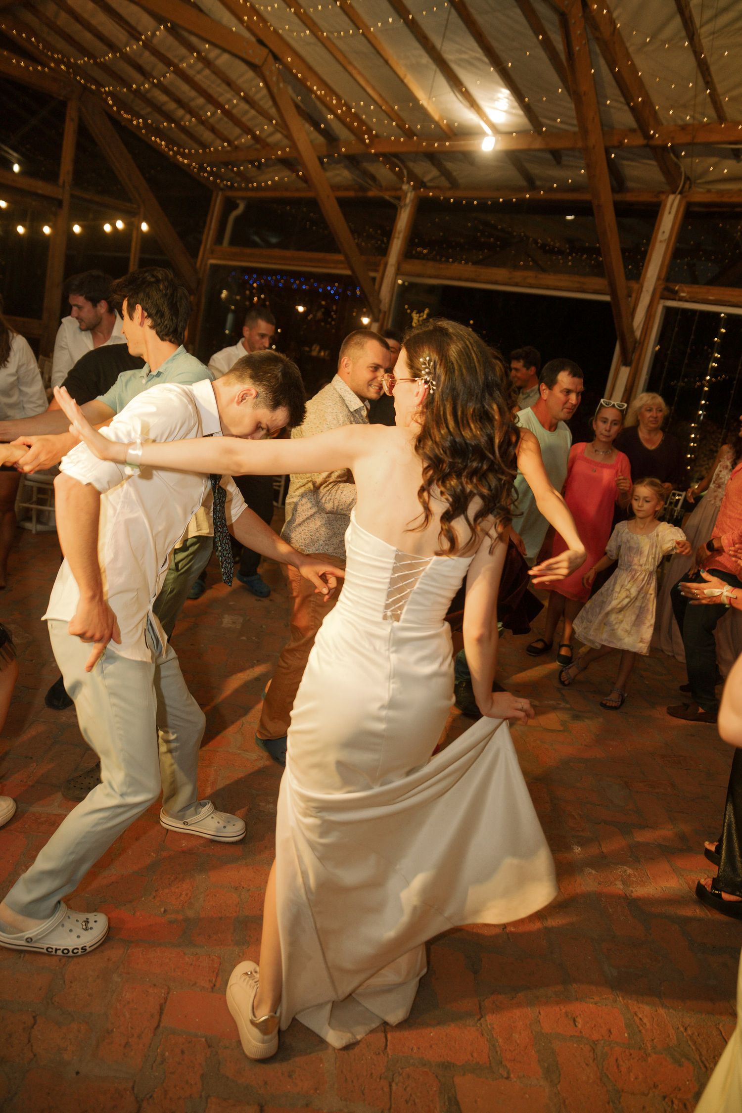Wedding guests dance and celebrate on a rustic brick floor under twinkling string lights at an indoor reception venue.