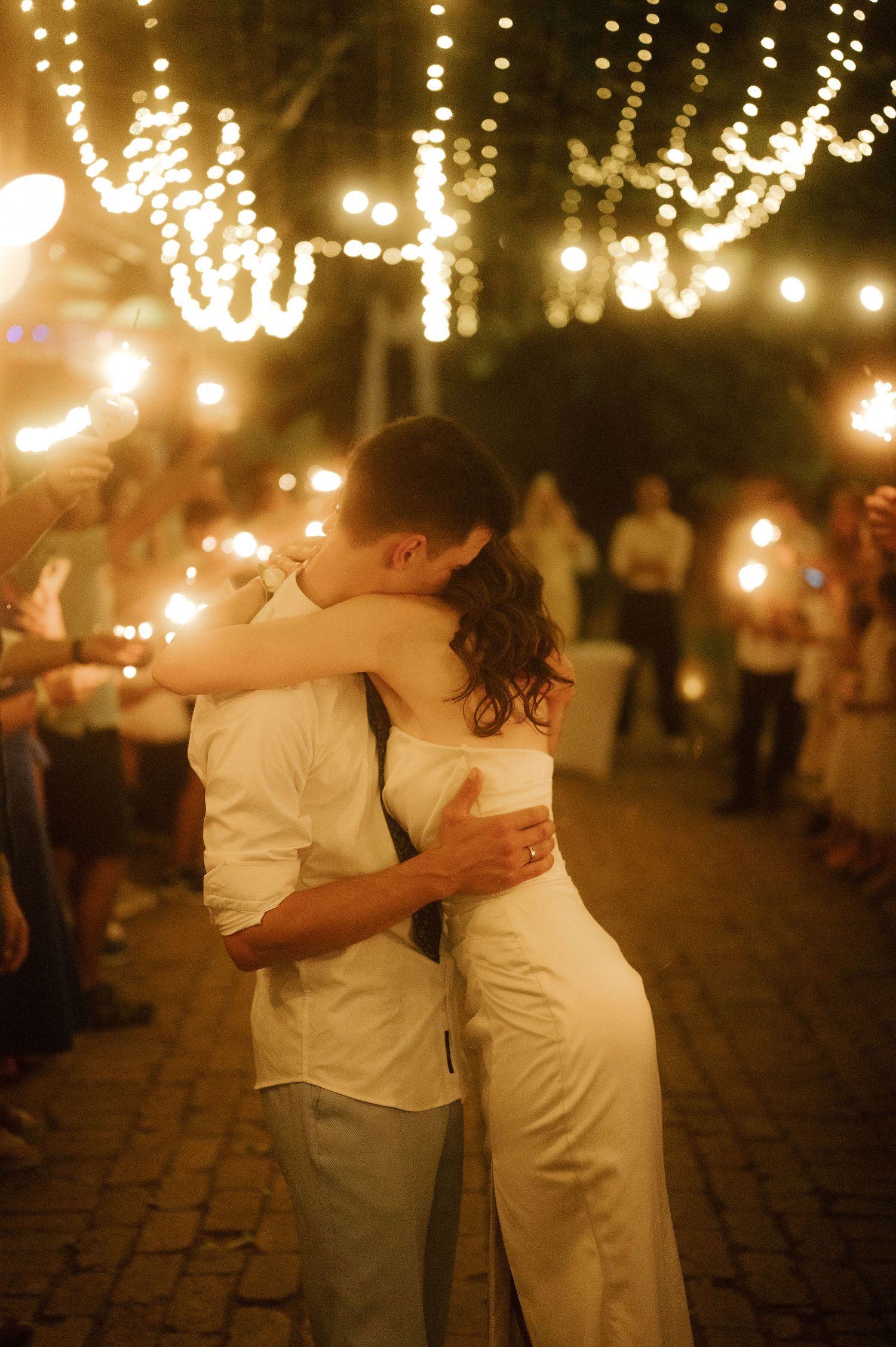 A romantic couple in white outfits dances together under twinkling lights at an evening outdoor celebration.