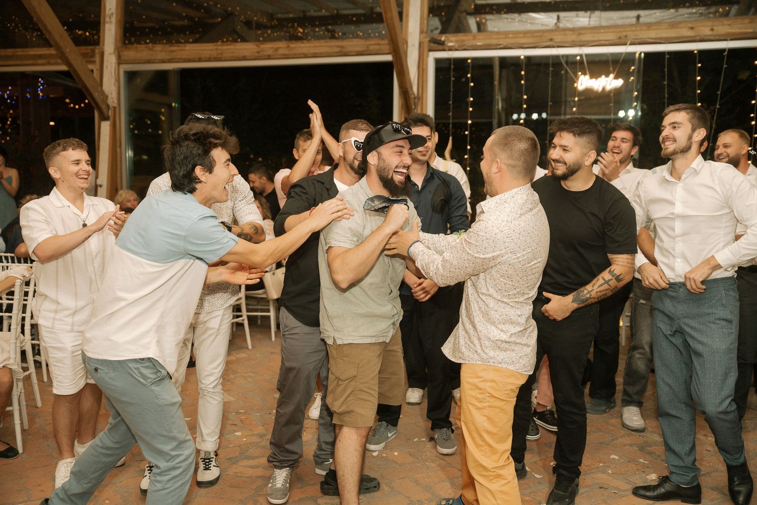 Wedding guests form a lively dance line at a rustic barn reception venue, moving in synchronized celebration.