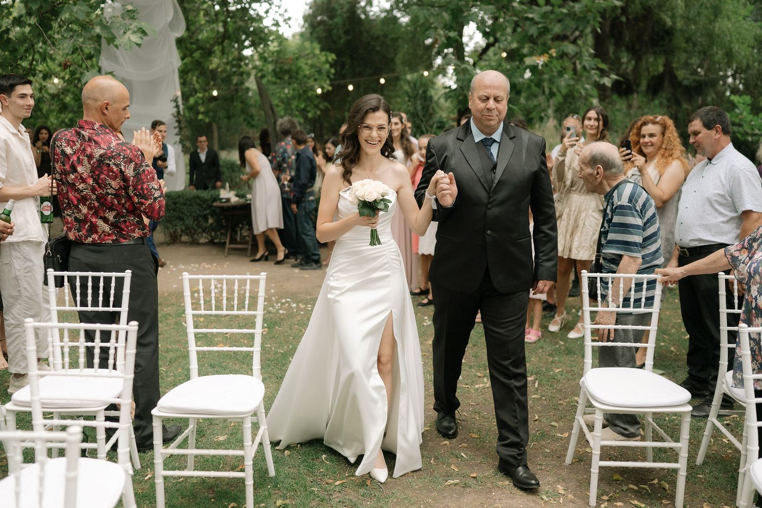 A panoramic view of an outdoor wedding ceremony setup with white chairs arranged in rows under green trees.