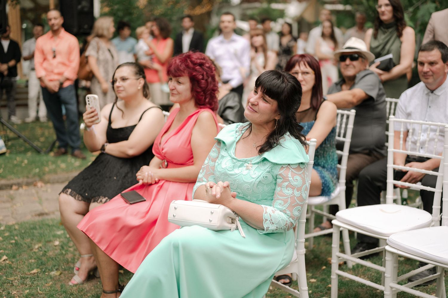 Wedding guests in vintage-style dresses sit on white chairs during an outdoor garden ceremony.