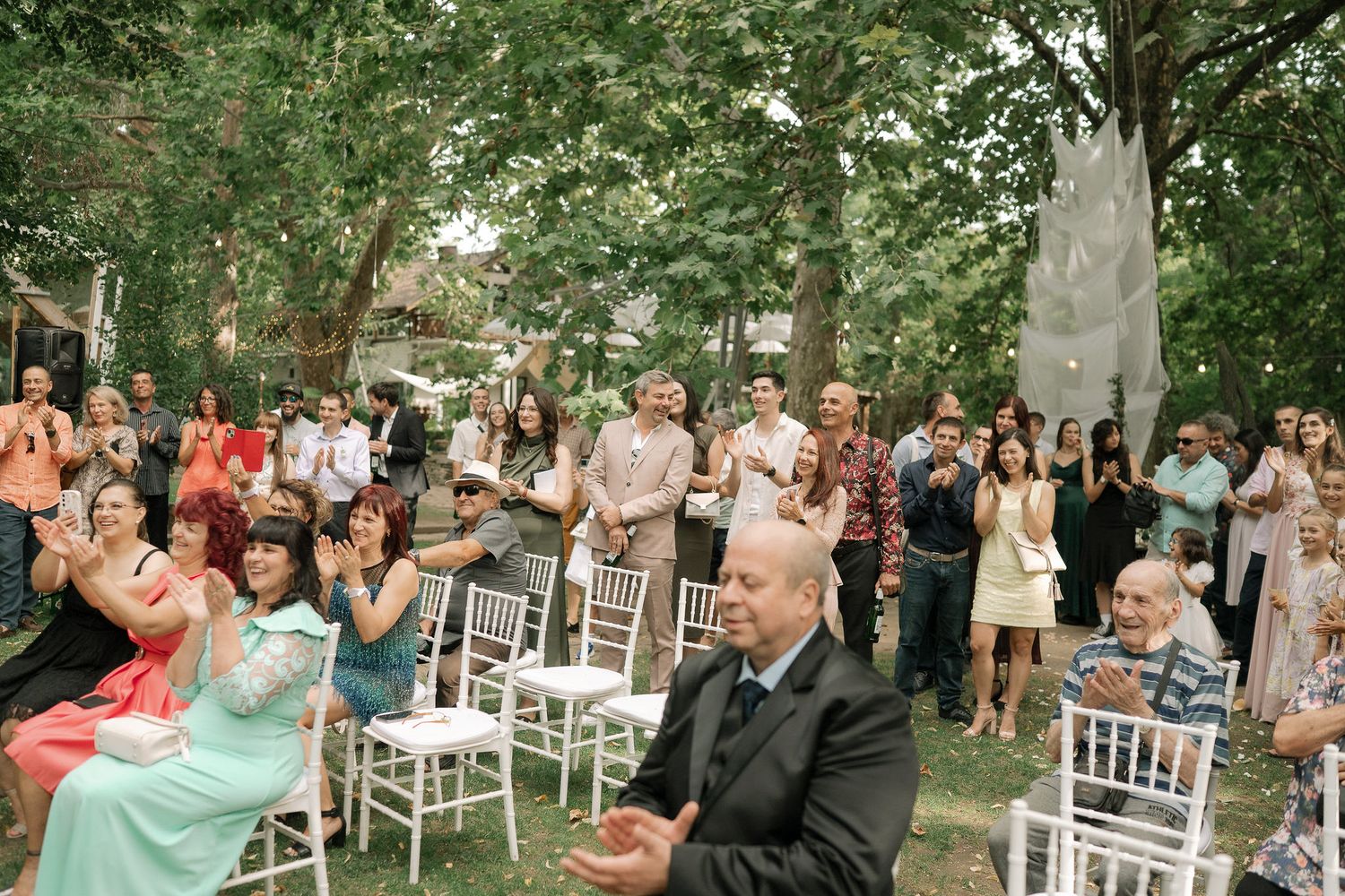 Outdoor garden wedding ceremony with white chairs arranged in rows surrounded by lush green trees and foliage.