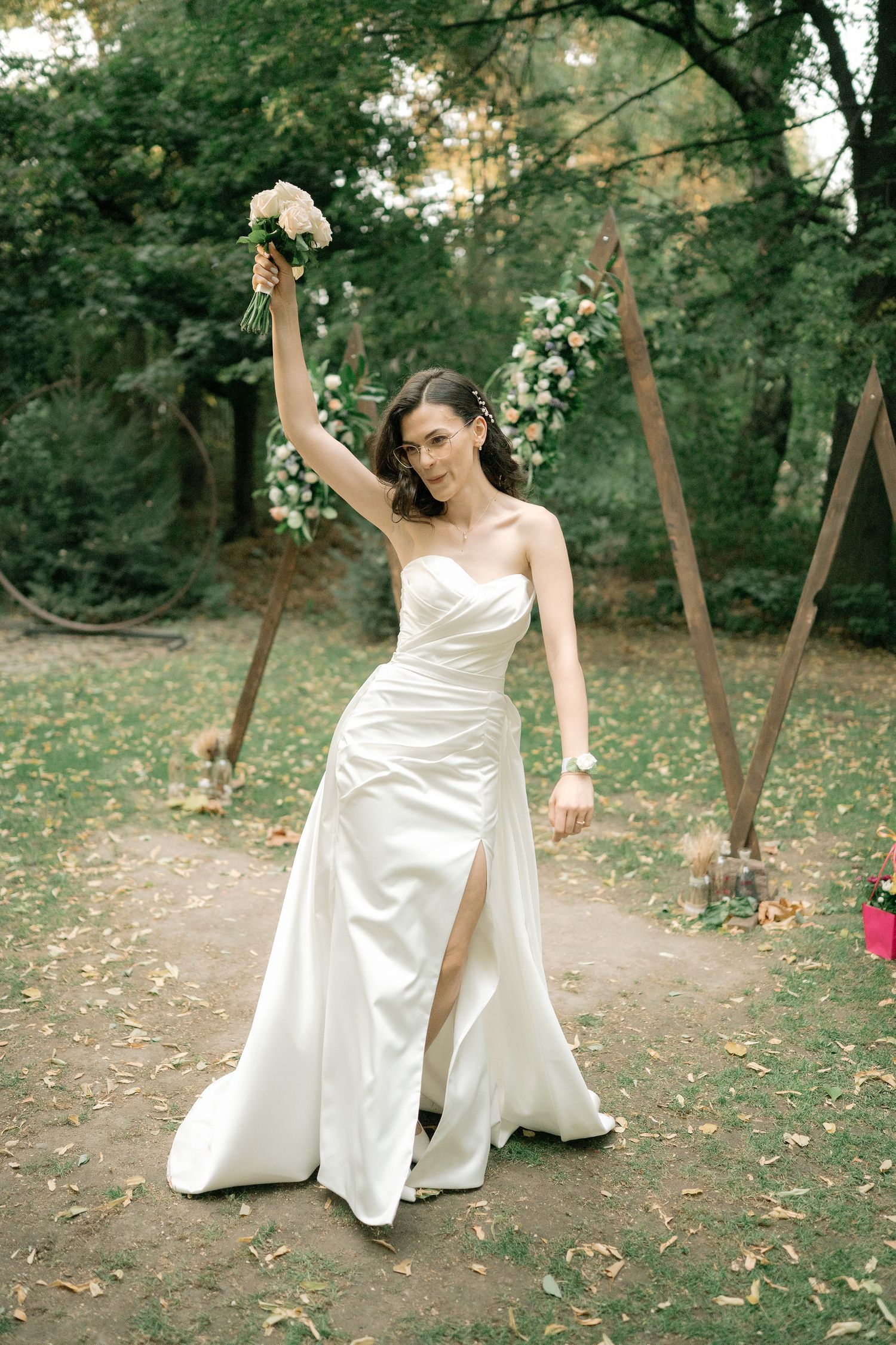 A bride in a flowing white satin wedding dress dances and twirls joyfully in a wooded outdoor setting.