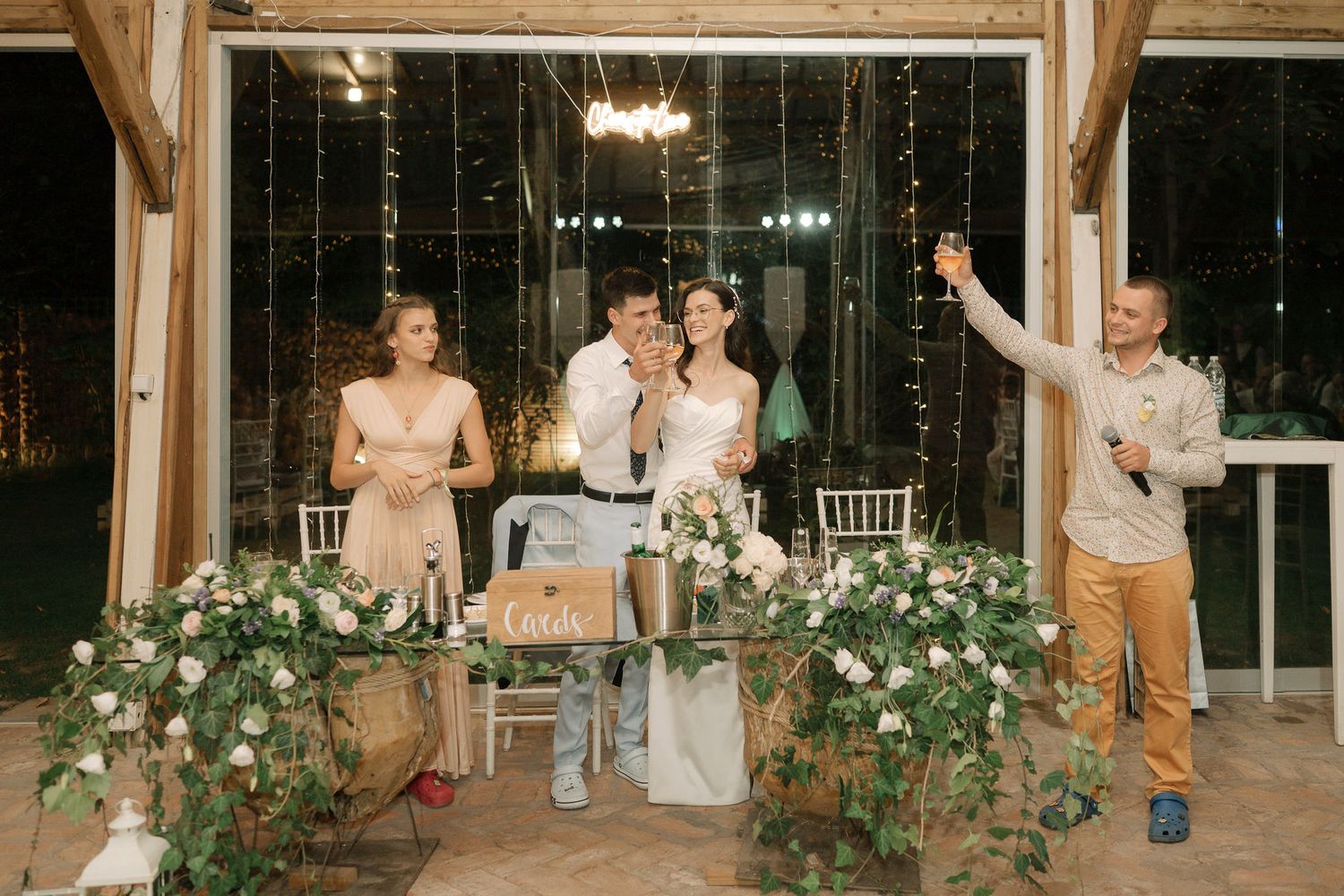 Wedding reception guests enjoy an evening celebration in a glass-enclosed venue with romantic lighting and floral decor.