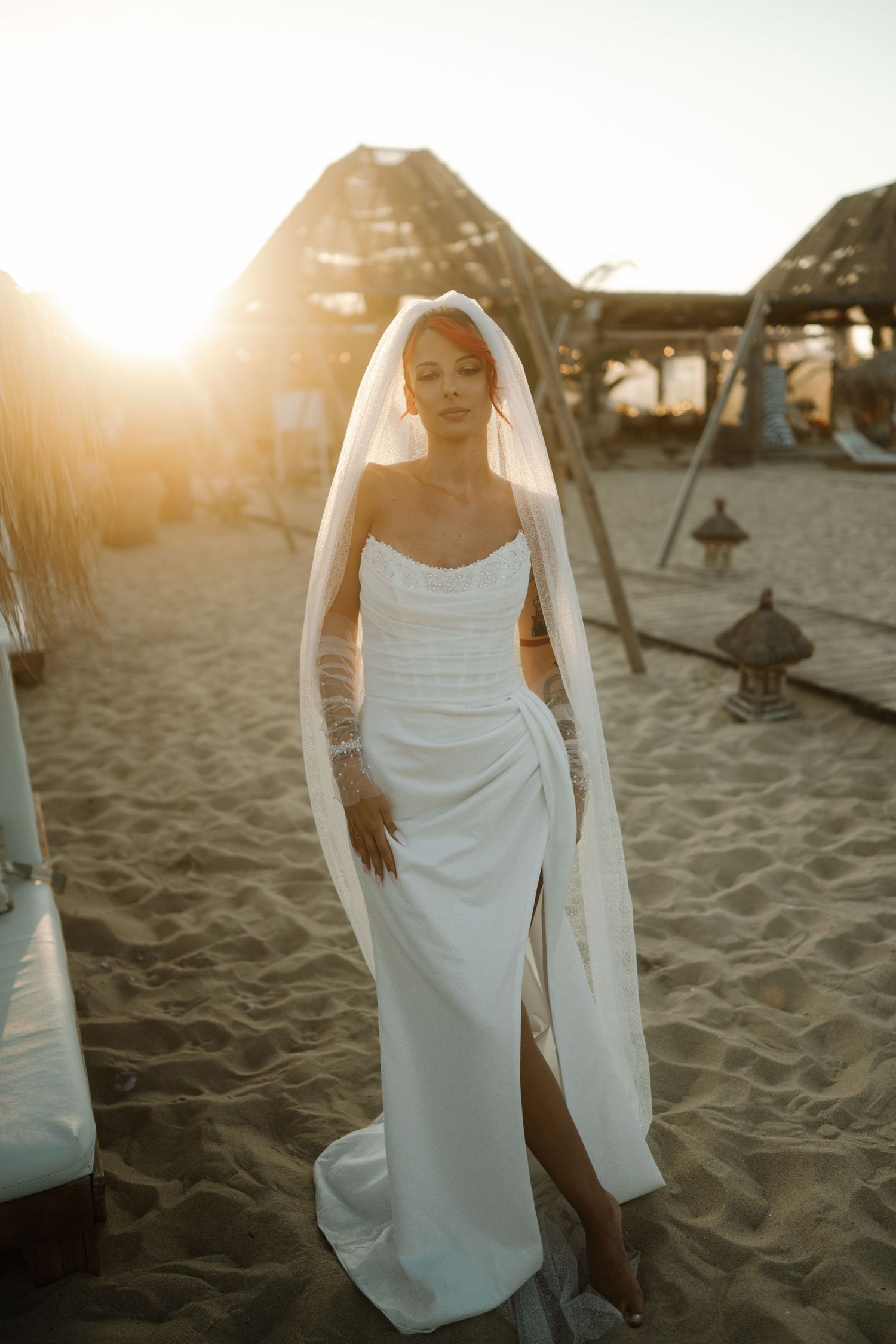A figure in a flowing white dress walks gracefully along a sandy beach at sunset with umbrellas in the background.
