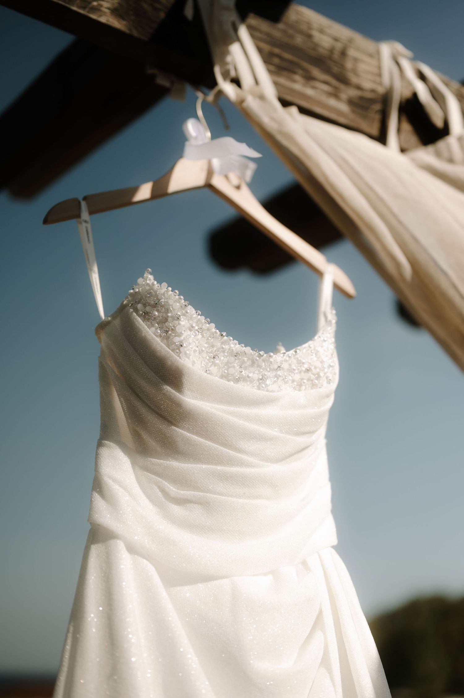 Close-up details of an elegant white wedding dress bodice with delicate beading and ruched fabric against a blue sky.