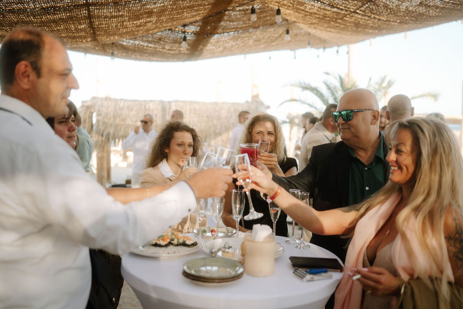 A group of people enjoying conversation and food at a rustic outdoor dining table under a covered patio.