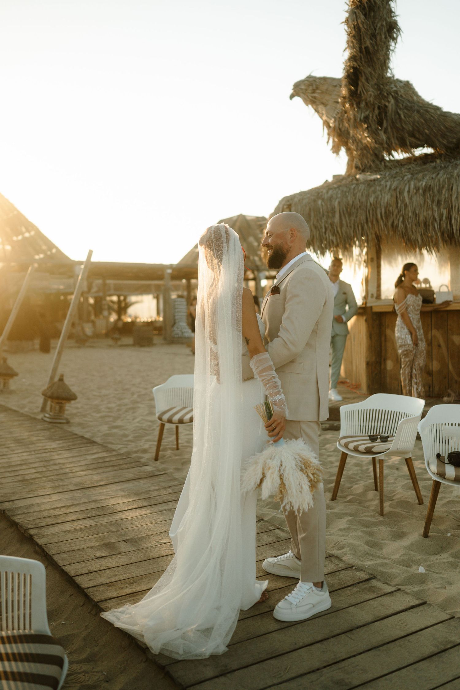 A romantic beach wedding ceremony at sunset with couple dancing on a wooden deck surrounded by thatched umbrellas.