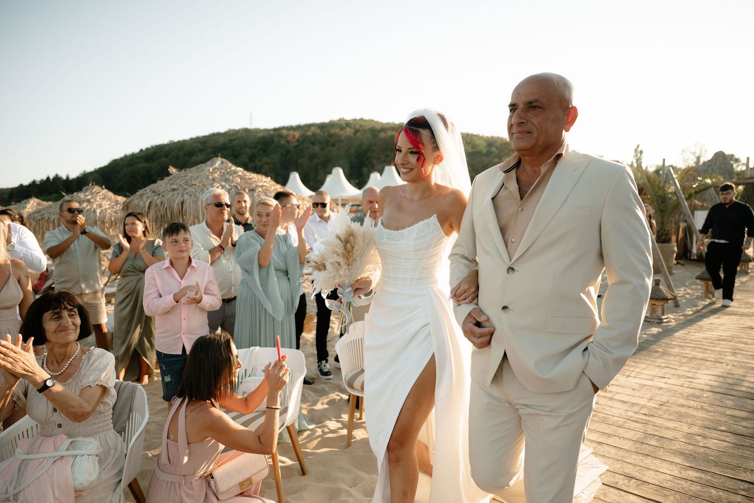 A joyful outdoor wedding celebration on a wooden deck with guests in light summer attire clapping for the newlyweds.