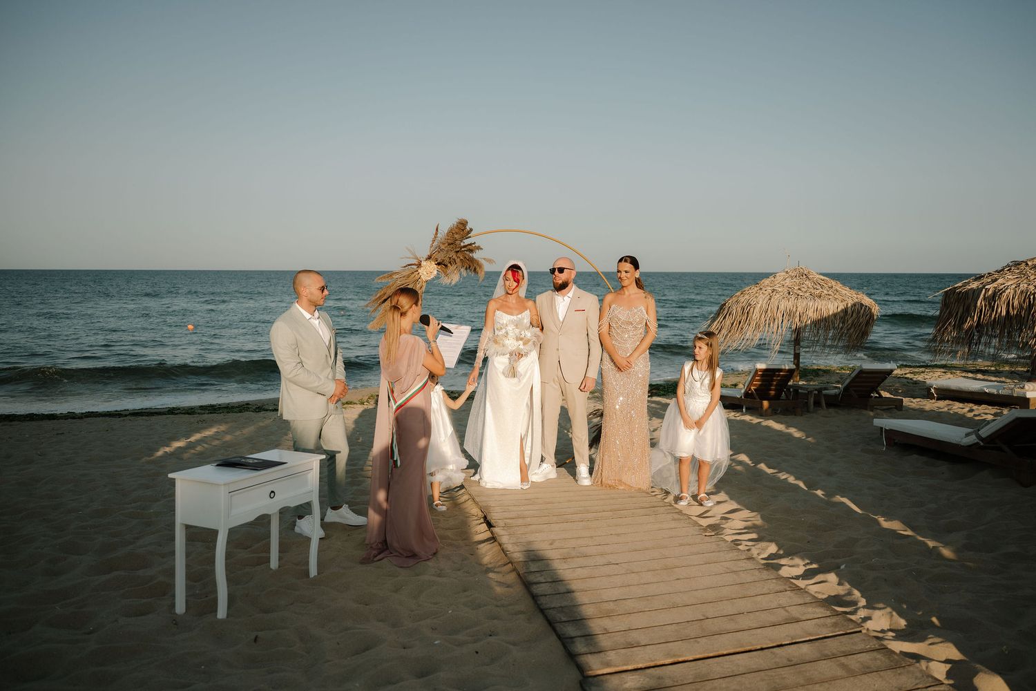 A wedding party stands on a wooden boardwalk at sunset by the ocean with a white piano and large rocks in the background.
