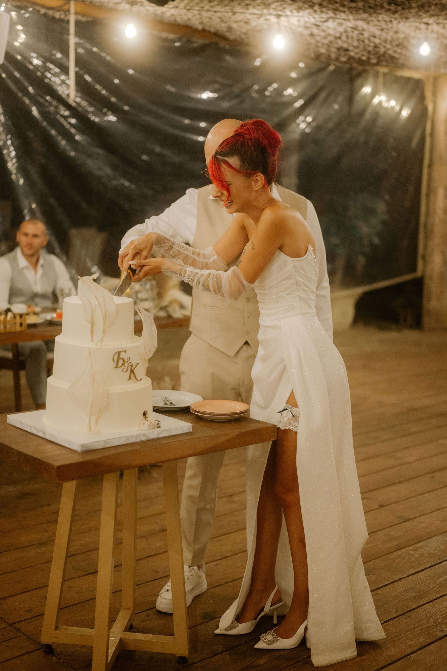 Wedding couple cuts their elegant white cake together at a rustic outdoor reception under string lights.