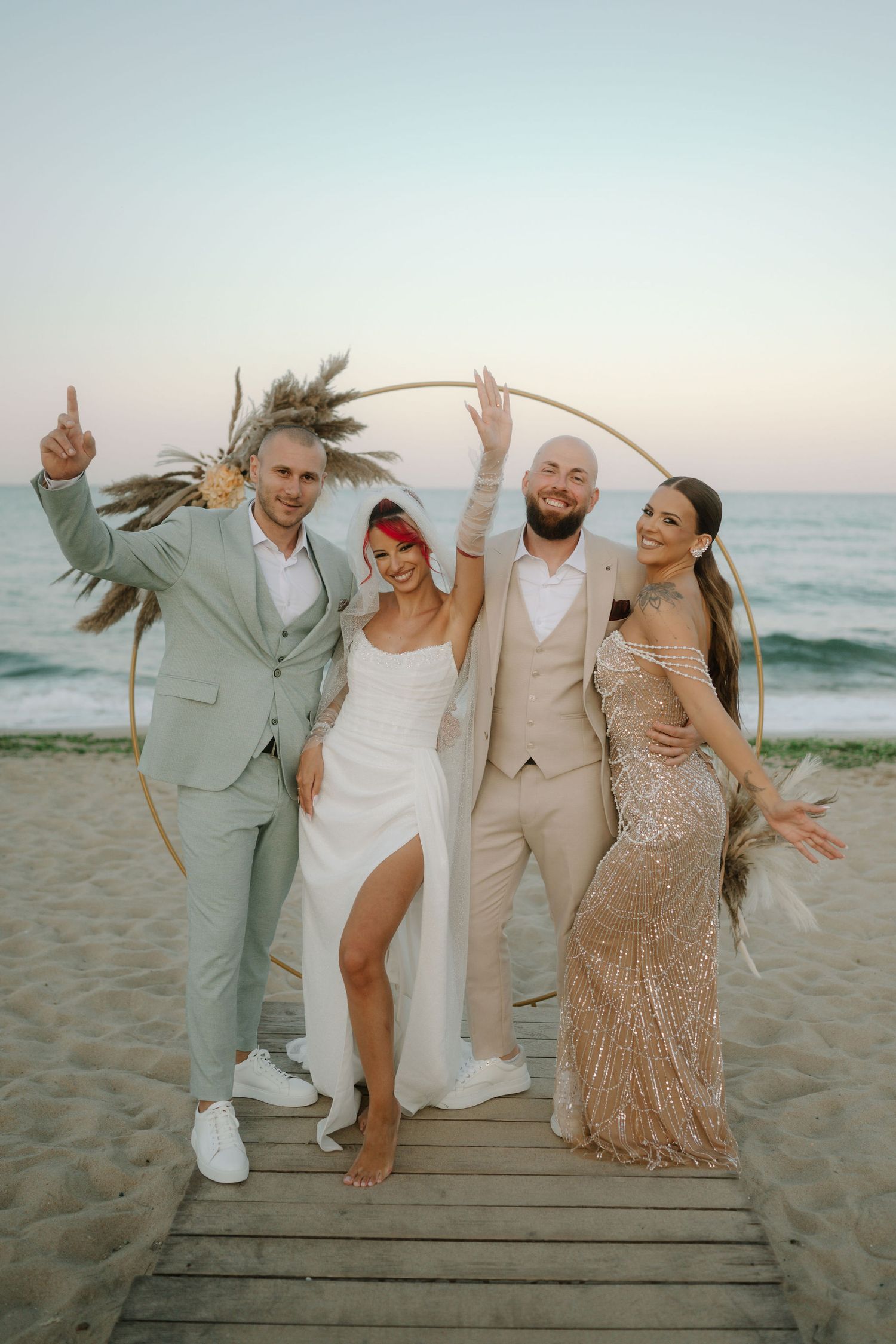 Wedding party celebrates joyfully while jumping on wooden boardwalk at sunset beach in coordinated beige attire.