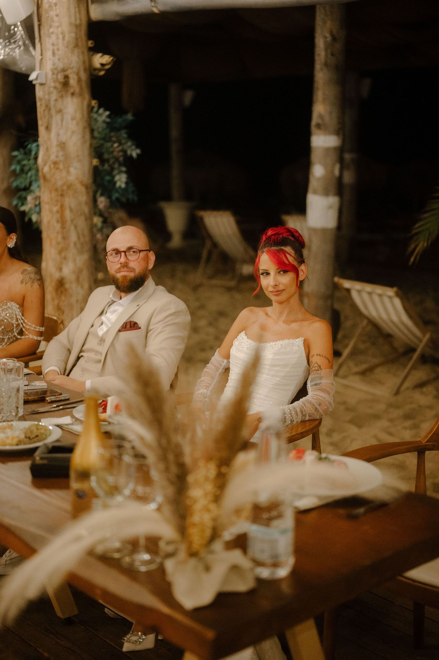 Couple seated at rustic outdoor wedding reception with dried pampas grass centerpiece and wooden decor at night.