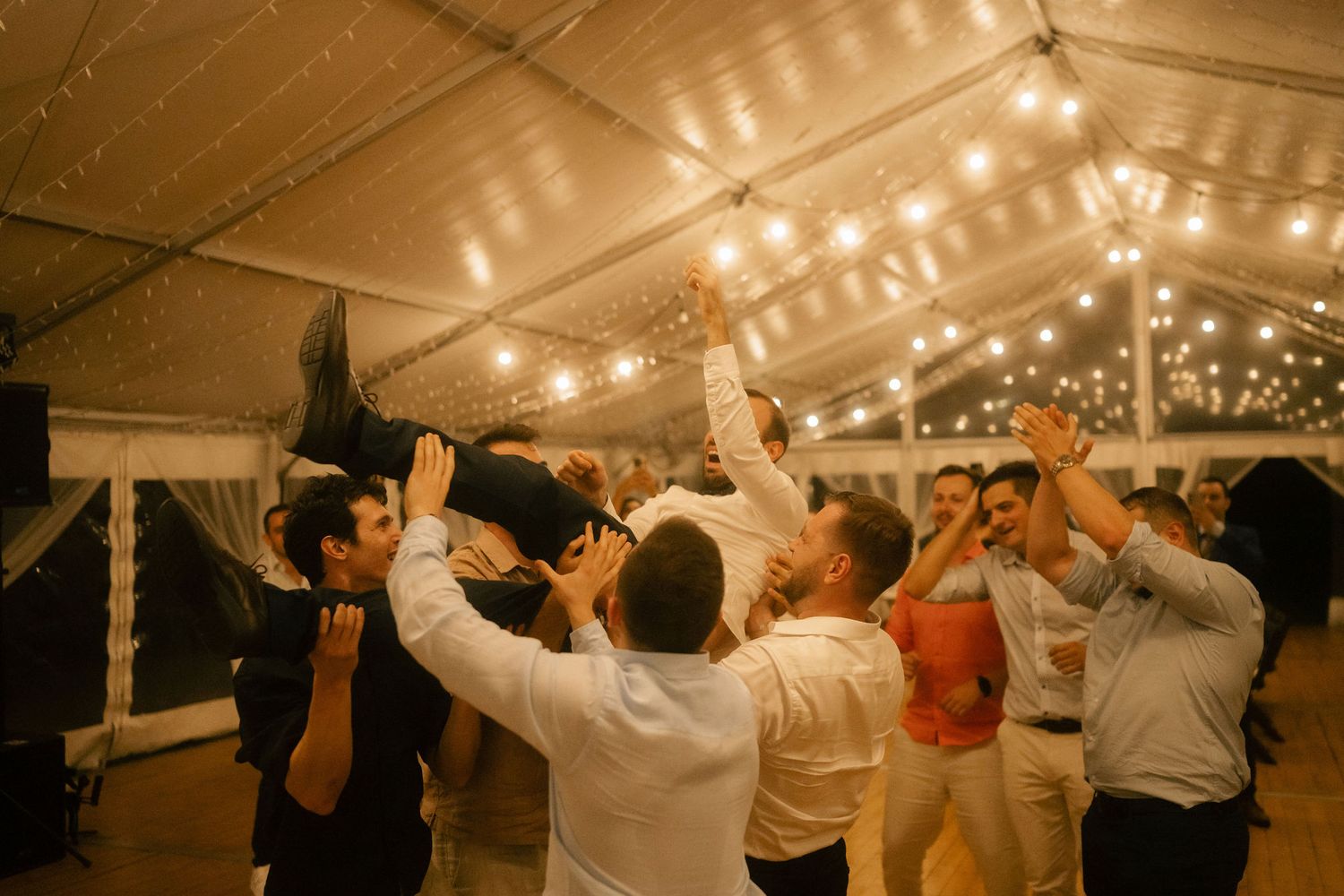Wedding guests dancing with raised arms under a glowing tent canopy at an evening reception celebration.