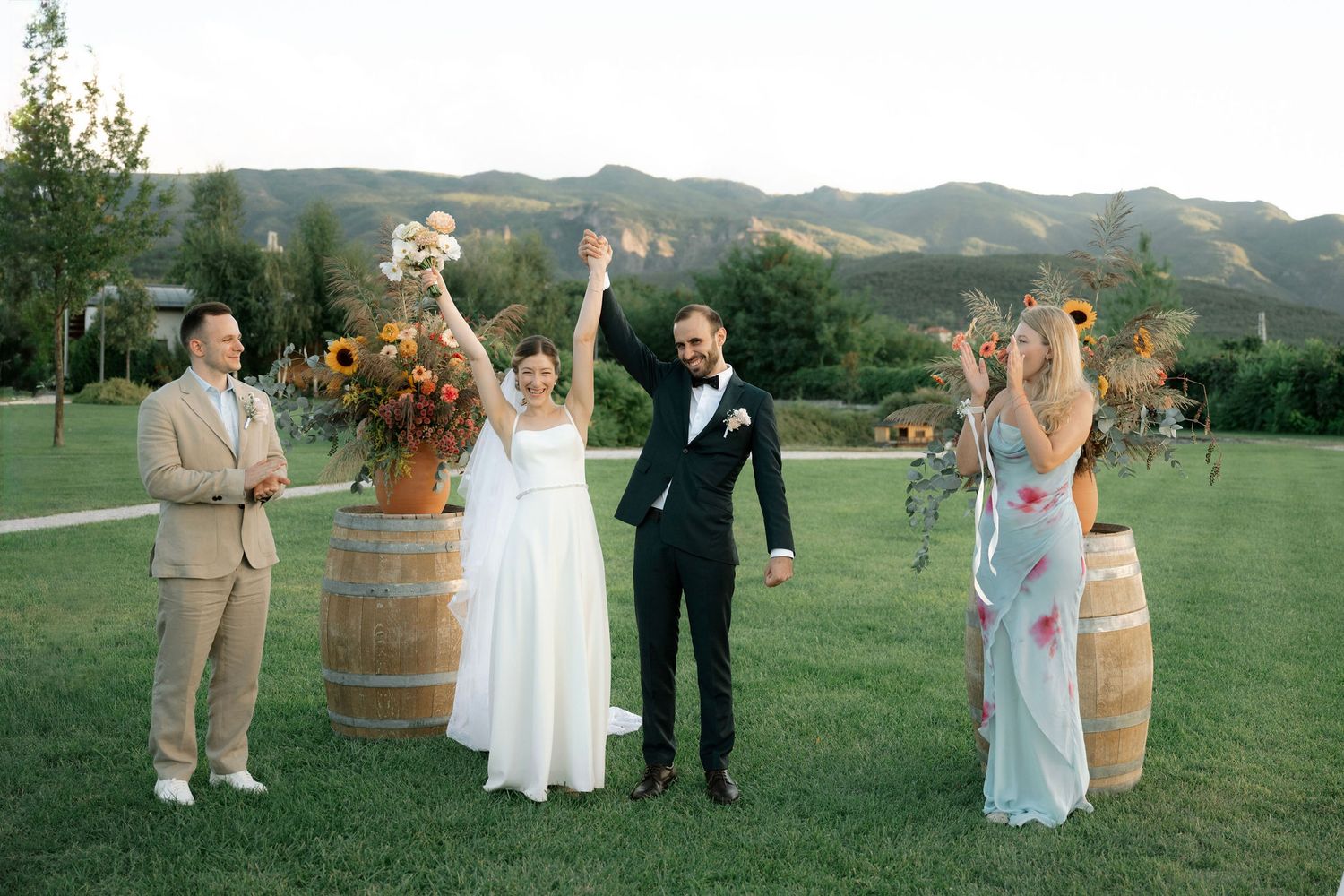 Wedding party poses in a scenic mountain vineyard setting with rustic wine barrels and rolling hills in background.
