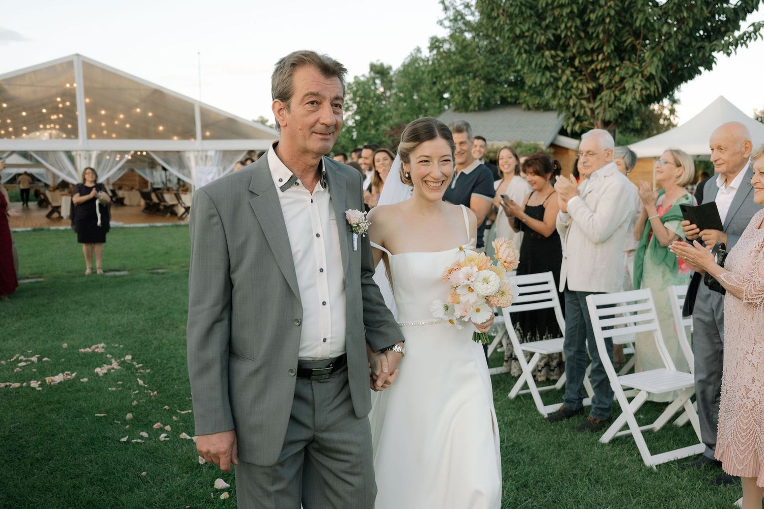 A wedding celebration in an outdoor garden setting with guests seated in white chairs during a summer evening.