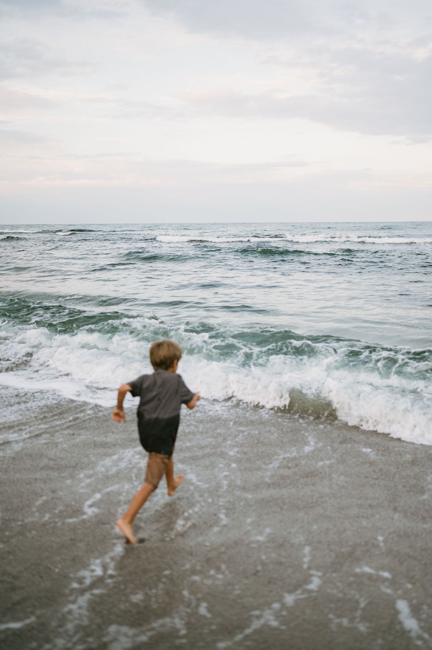 A young boy runs barefoot toward ocean waves on a sandy beach at dusk.