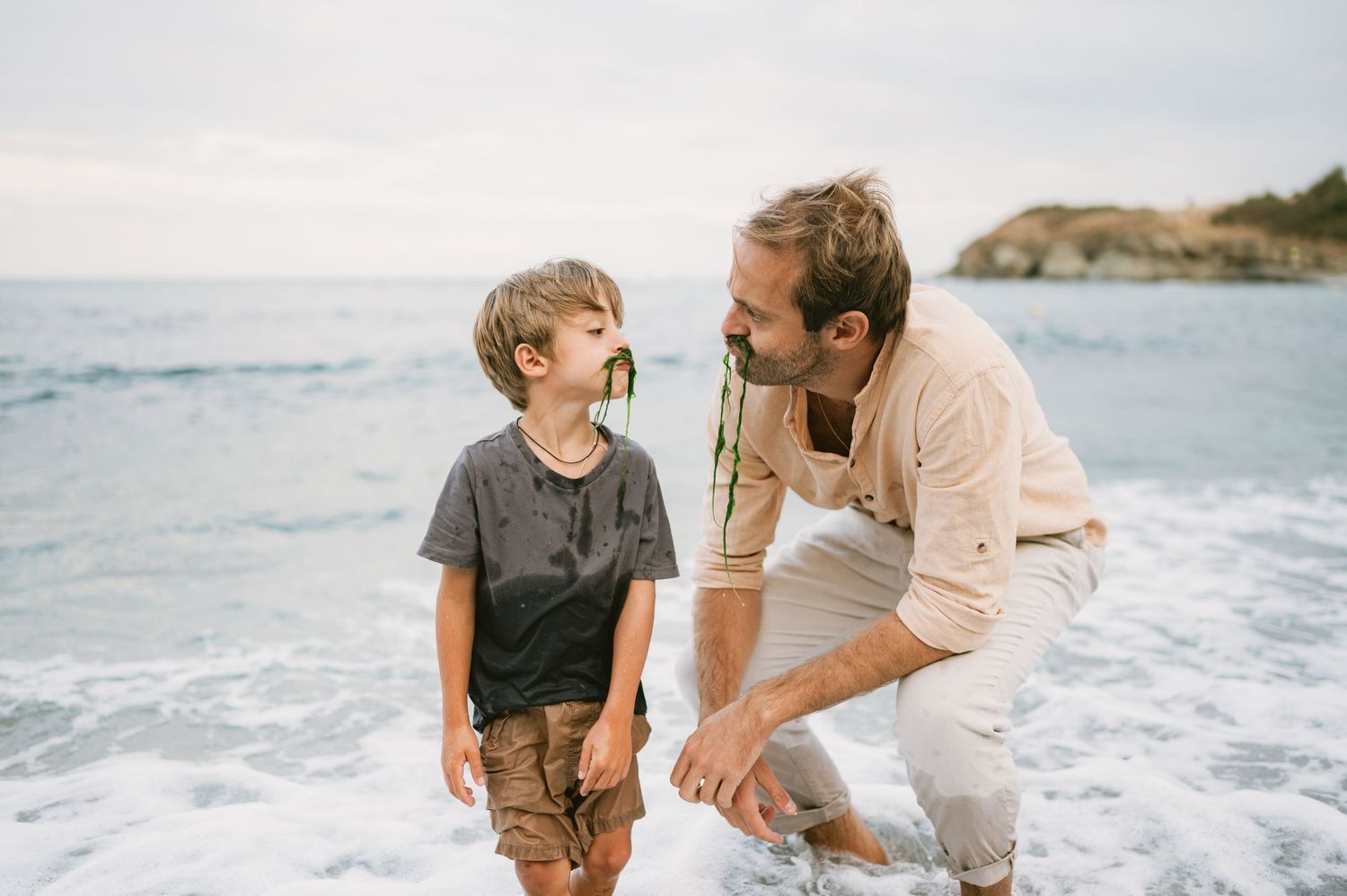 A father crouches down to talk with his young son on a scenic beach with ocean waves in the background.