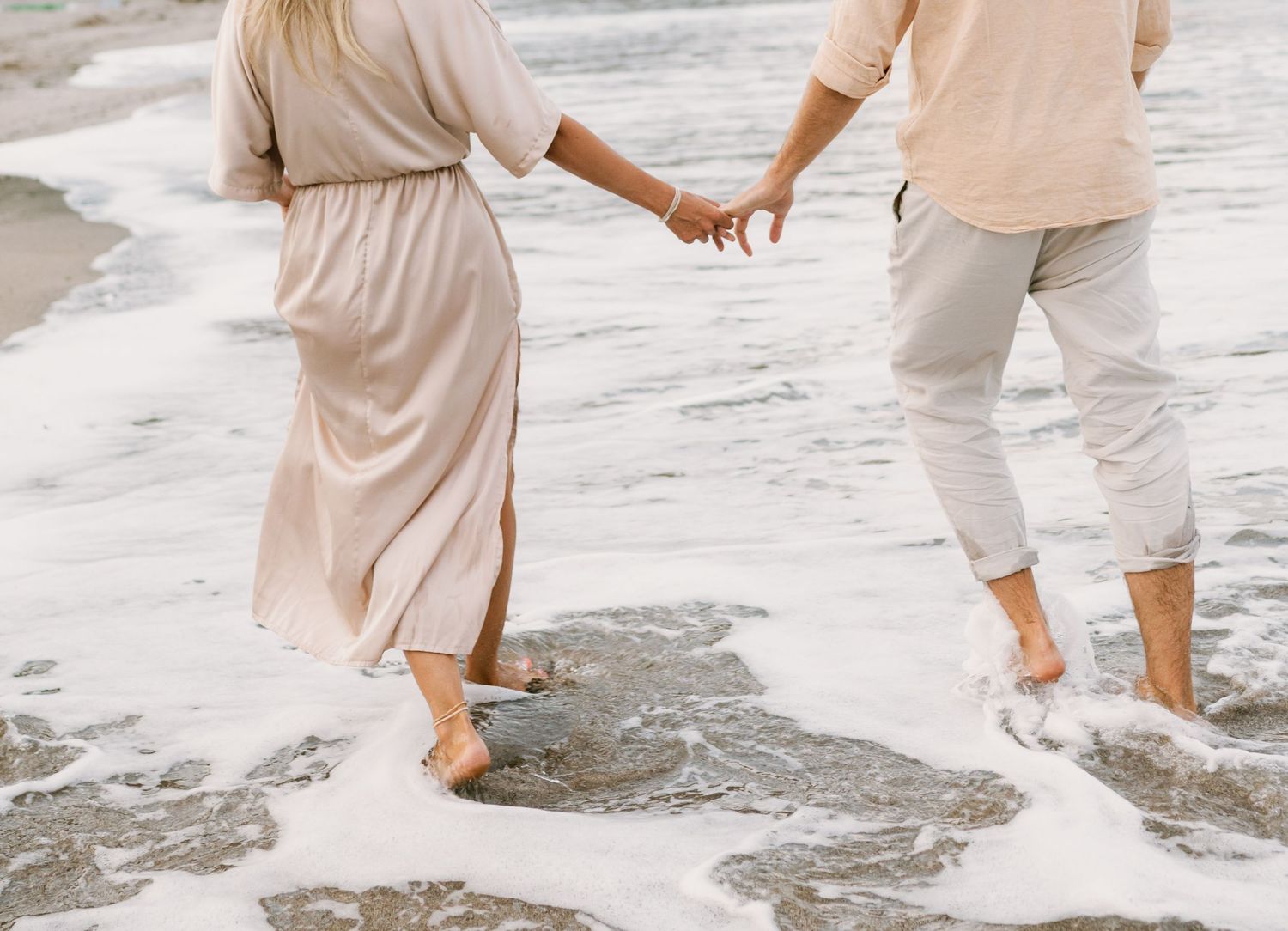Couple holding hands walking barefoot on a sandy beach with ocean waves washing over their feet.