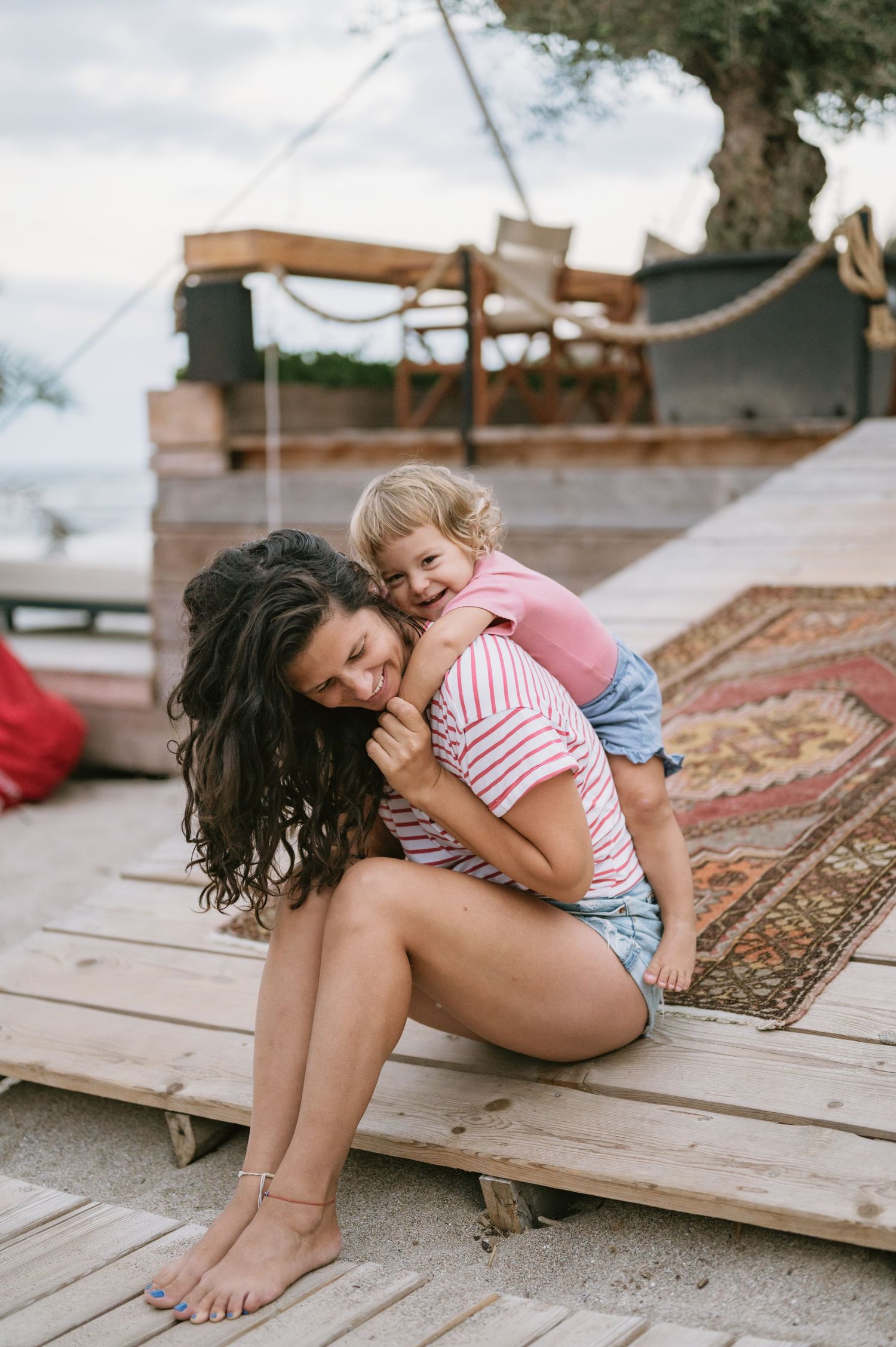 A mother sits on wooden decking while her laughing toddler hugs her from behind at an outdoor beach resort.