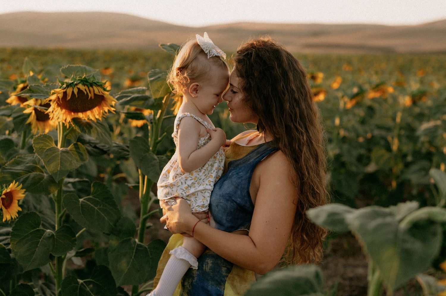A mother holds her baby girl in a sunflower field at golden hour, sharing a tender nose-to-nose moment.