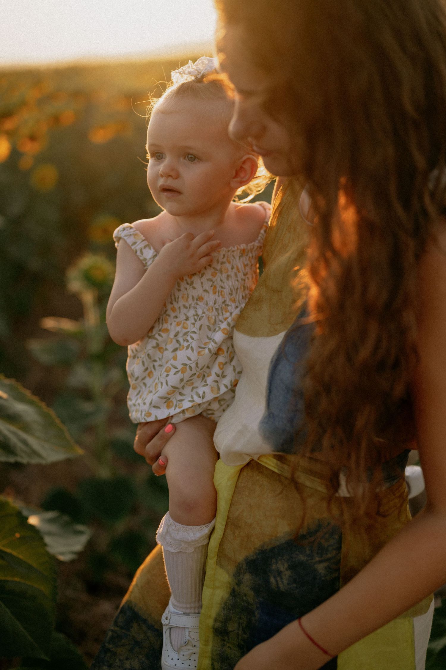 Mother holding baby girl in sunflower field at golden hour sunset, infant wearing floral romper and white socks.