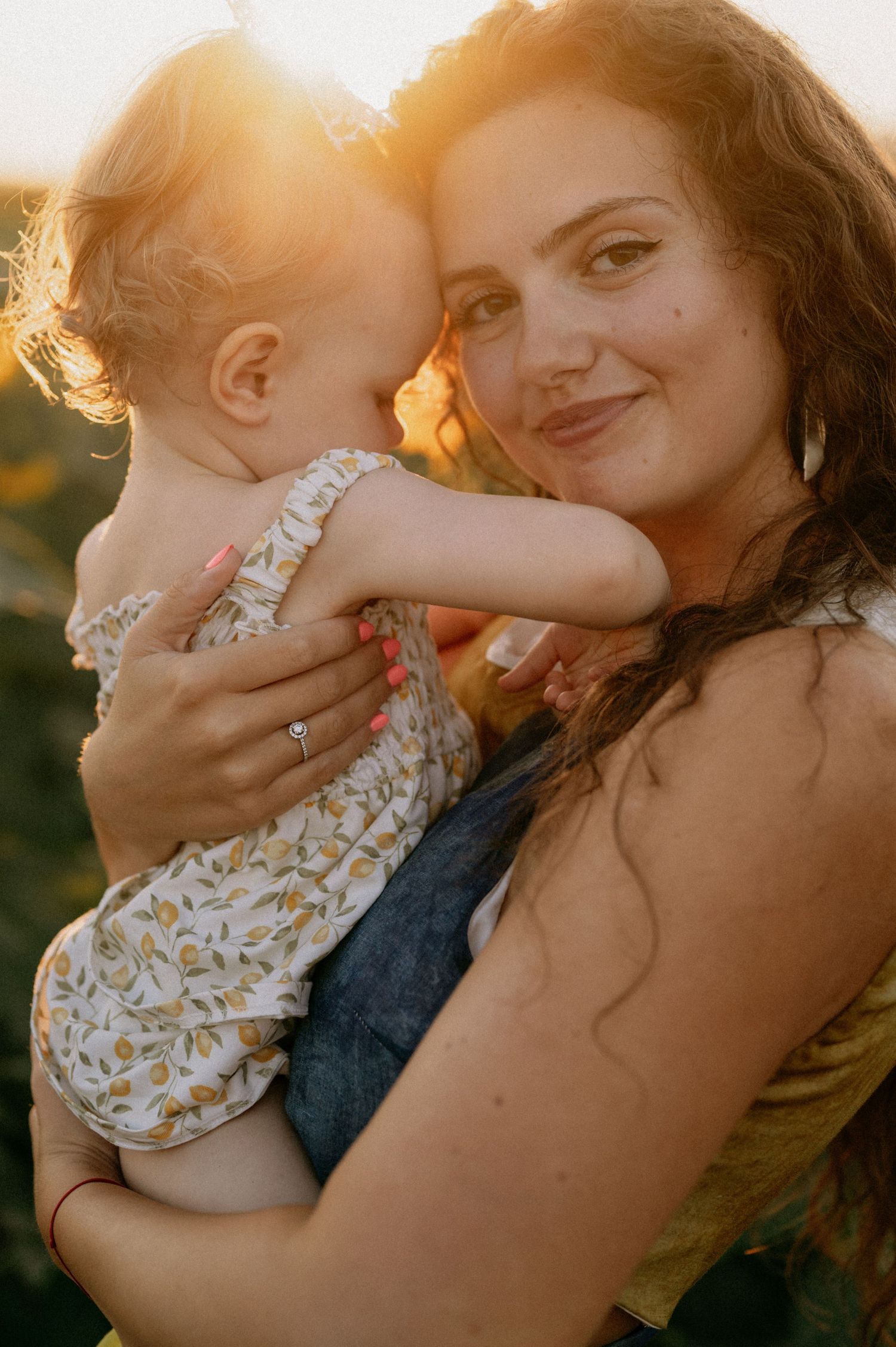 A smiling mother with curly hair holds her baby in a floral outfit during a golden sunset outdoors.