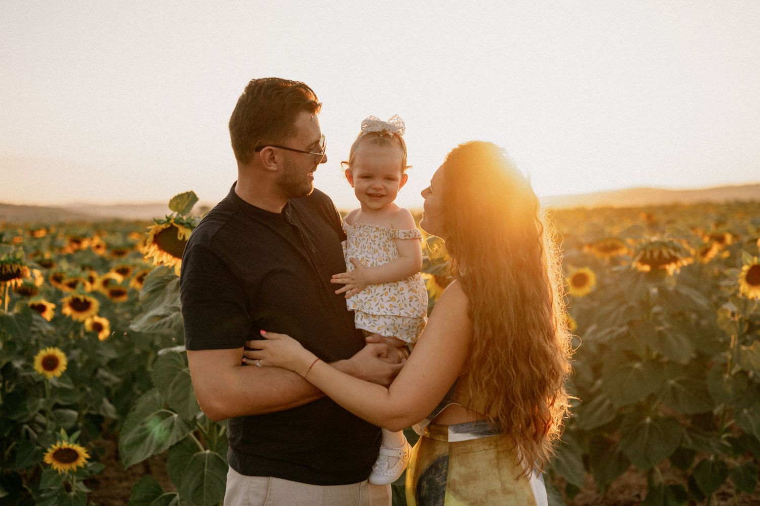 Family of three posing in a sunflower field at golden hour, parents holding toddler girl in white dress.