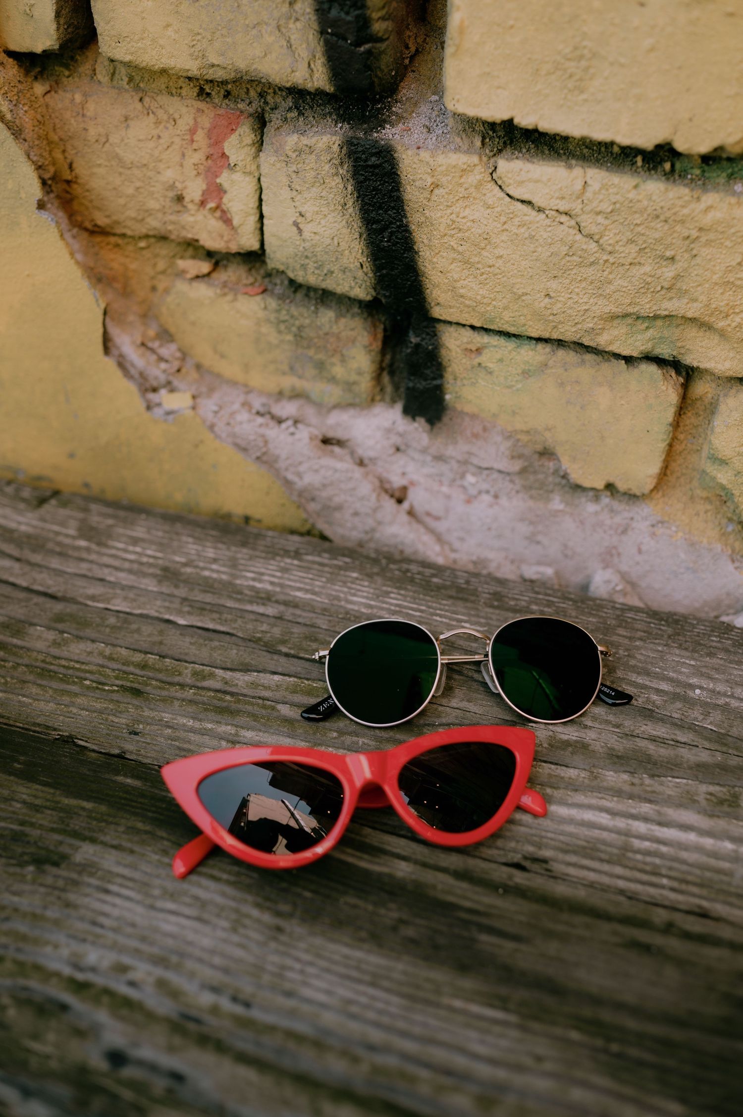 Two pairs of sunglasses, red cat-eye and round green lens, resting on weathered wood against a brick wall.