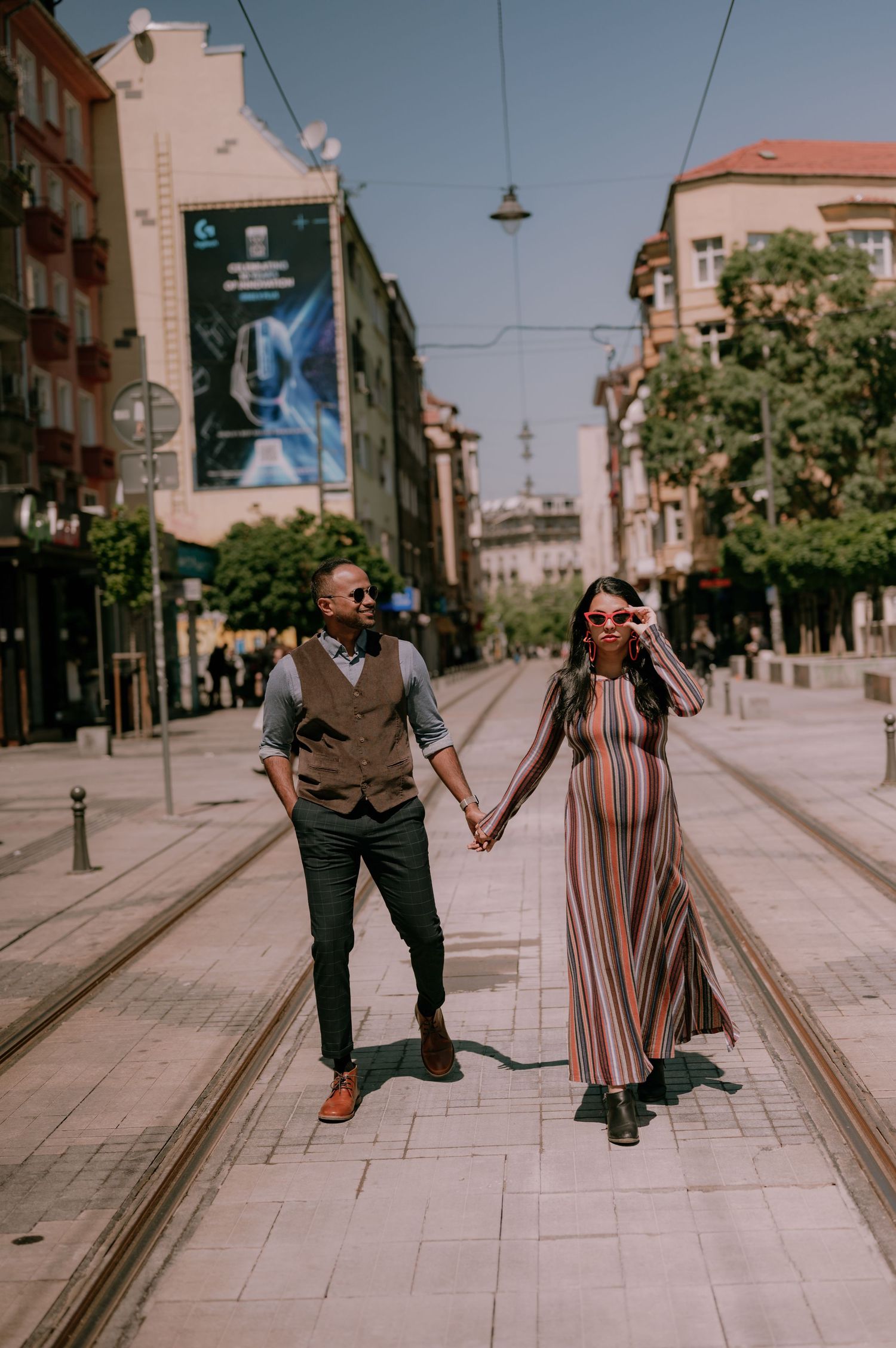 Stylish couple holding hands on a city street with tram tracks, man in vest and woman in striped maxi dress.