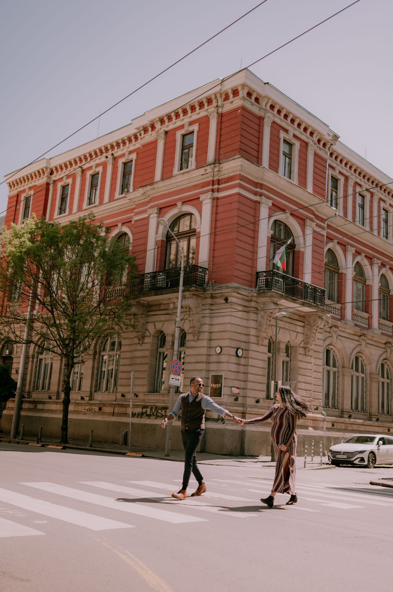 Couple holding hands crossing a zebra crossing in front of a red historic building on a sunny day.