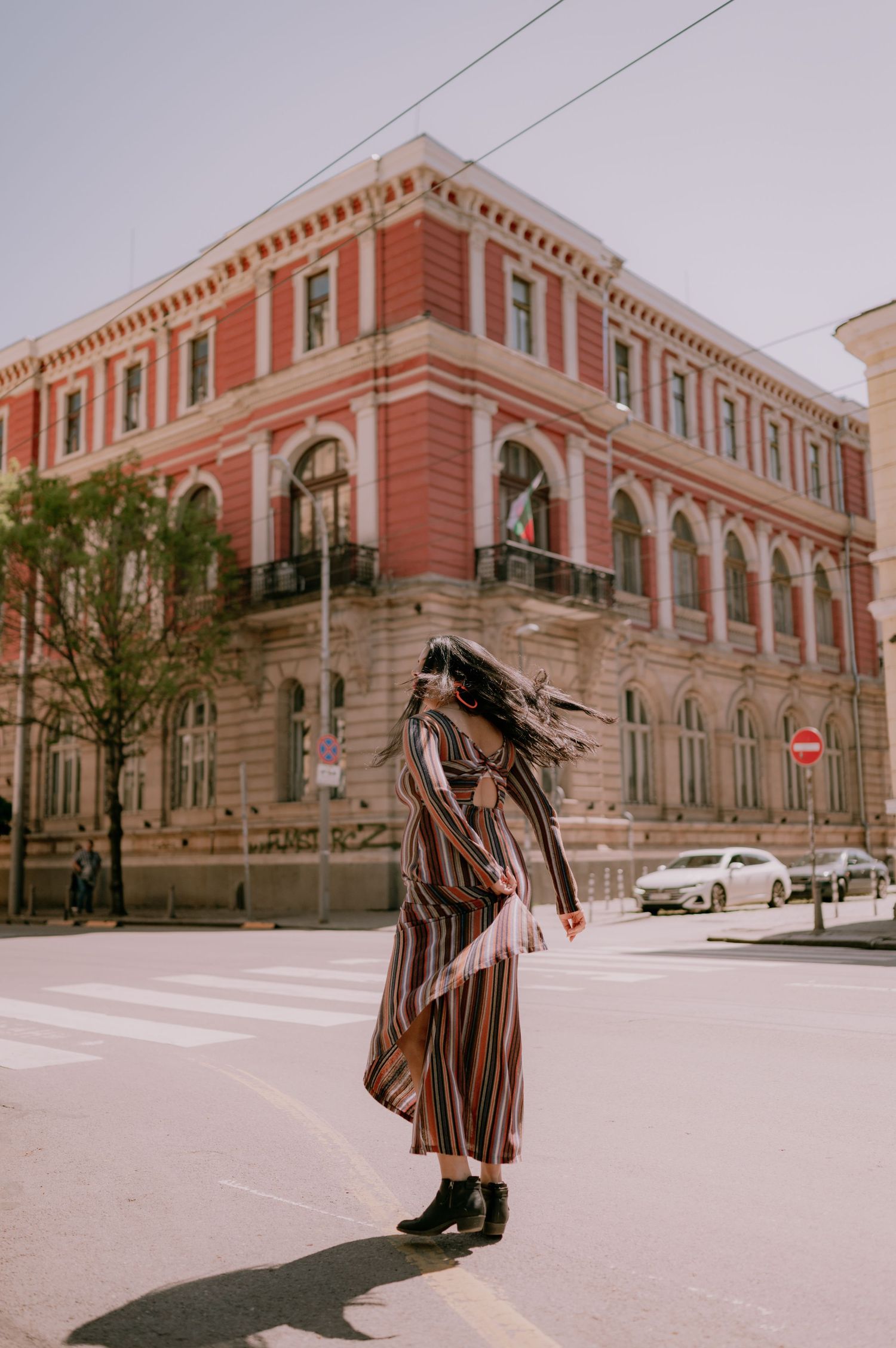Woman in striped maxi dress twirling on a sunny street corner near a grand red brick historic building.