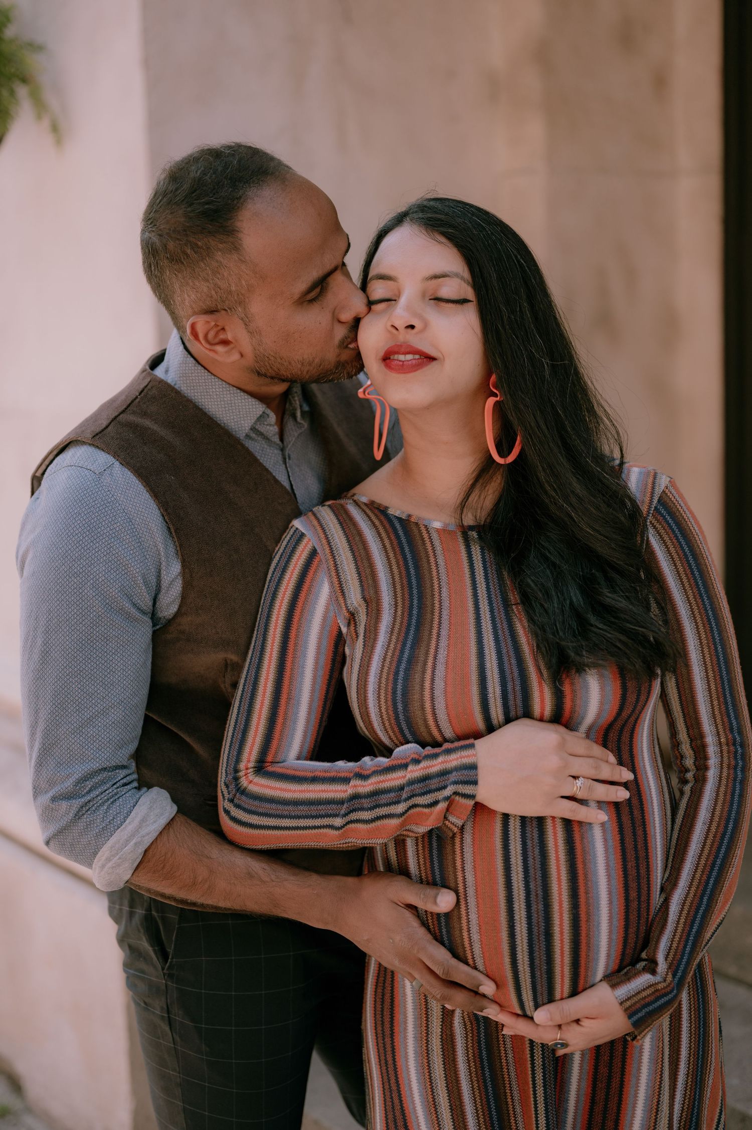 A couple shares a tender maternity moment, man kissing woman's temple while cradling her baby bump in striped dress.