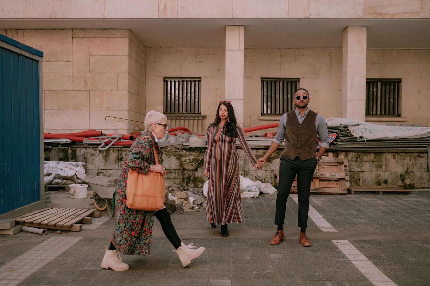 Three people walking outdoors near a stone building, woman in floral dress holding hands with companions.
