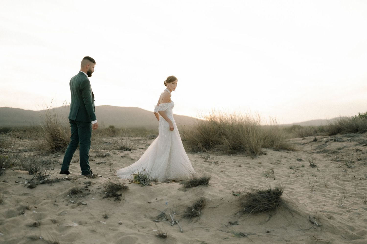 Bride and groom walking on sandy desert dunes at sunset, bride in elegant white gown with train.