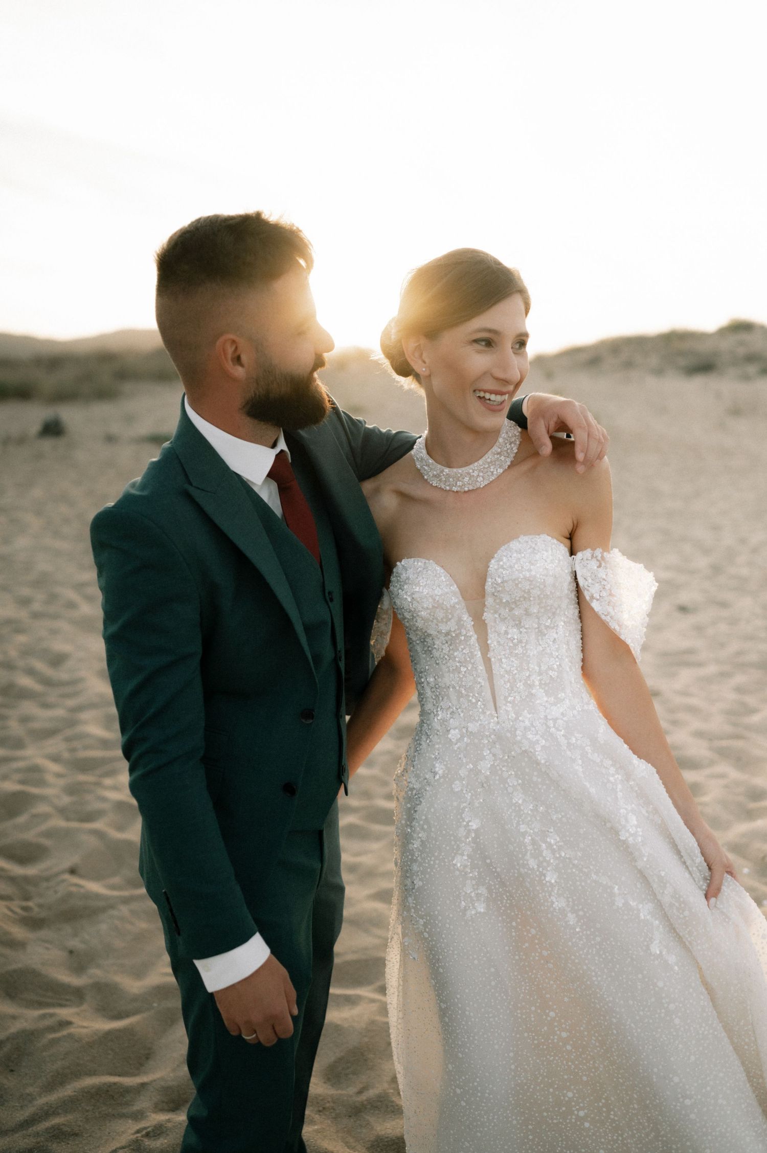 Smiling bride in sparkling off-shoulder gown and groom in teal suit pose on sandy beach at golden hour.