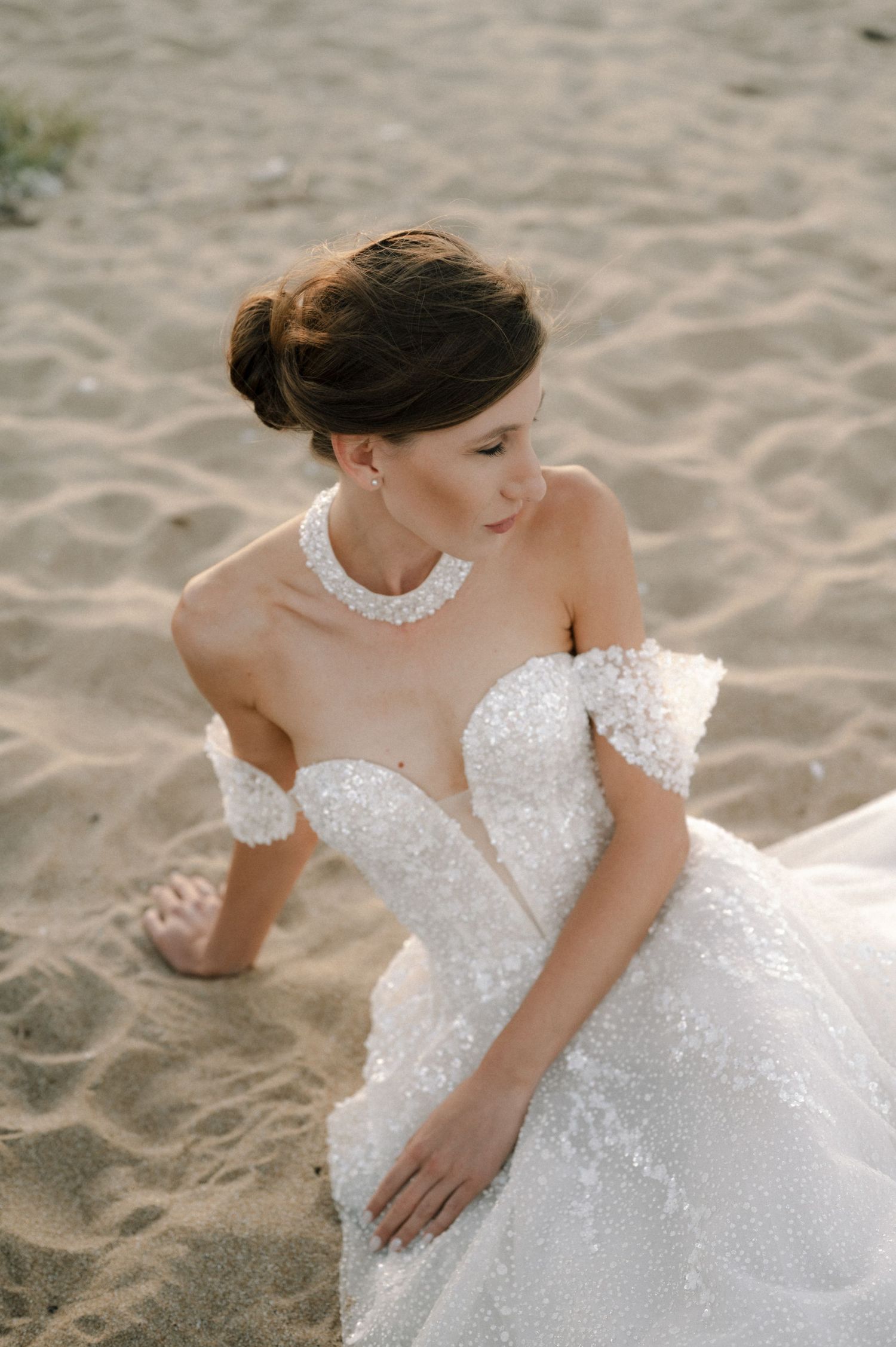 Elegant bride in off-shoulder beaded gown with pearl necklace sitting on sandy beach, looking gracefully downward.