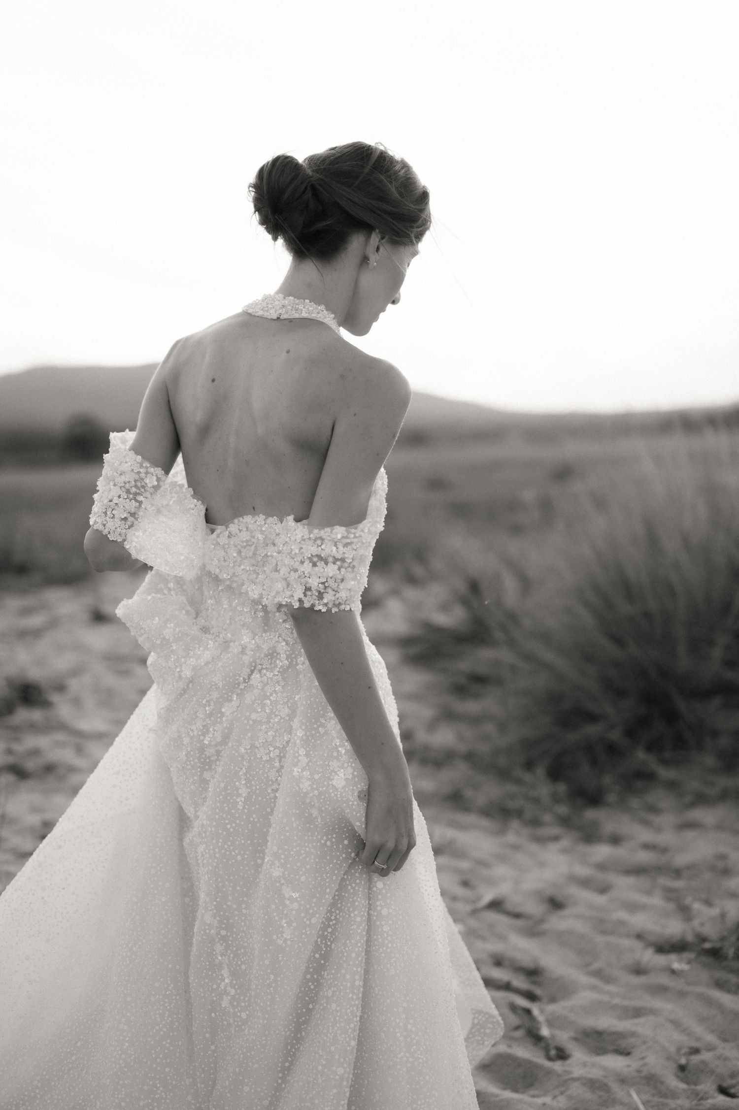 Elegant bride in a backless lace wedding gown with floral wrap poses in a sandy desert landscape, black and white photo.