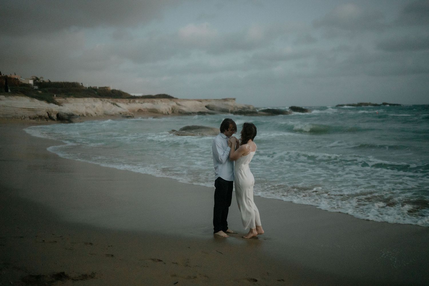 Couple embracing on a moody beach with rocky coastline and ocean waves in the background.