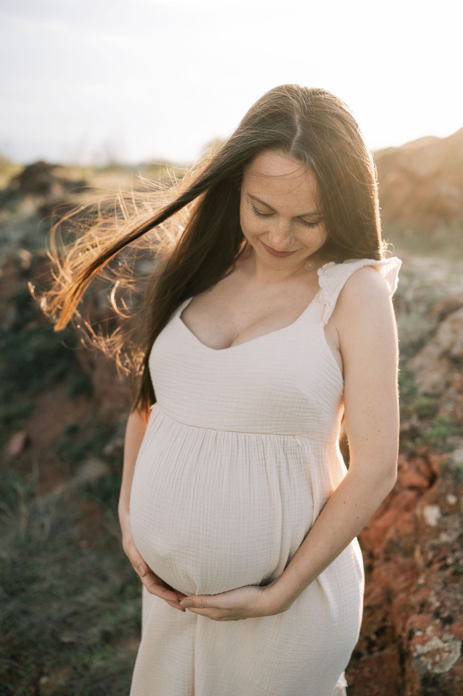 Expectant mother in flowing white dress poses outdoors during golden hour with sunlit hair and hands on belly.