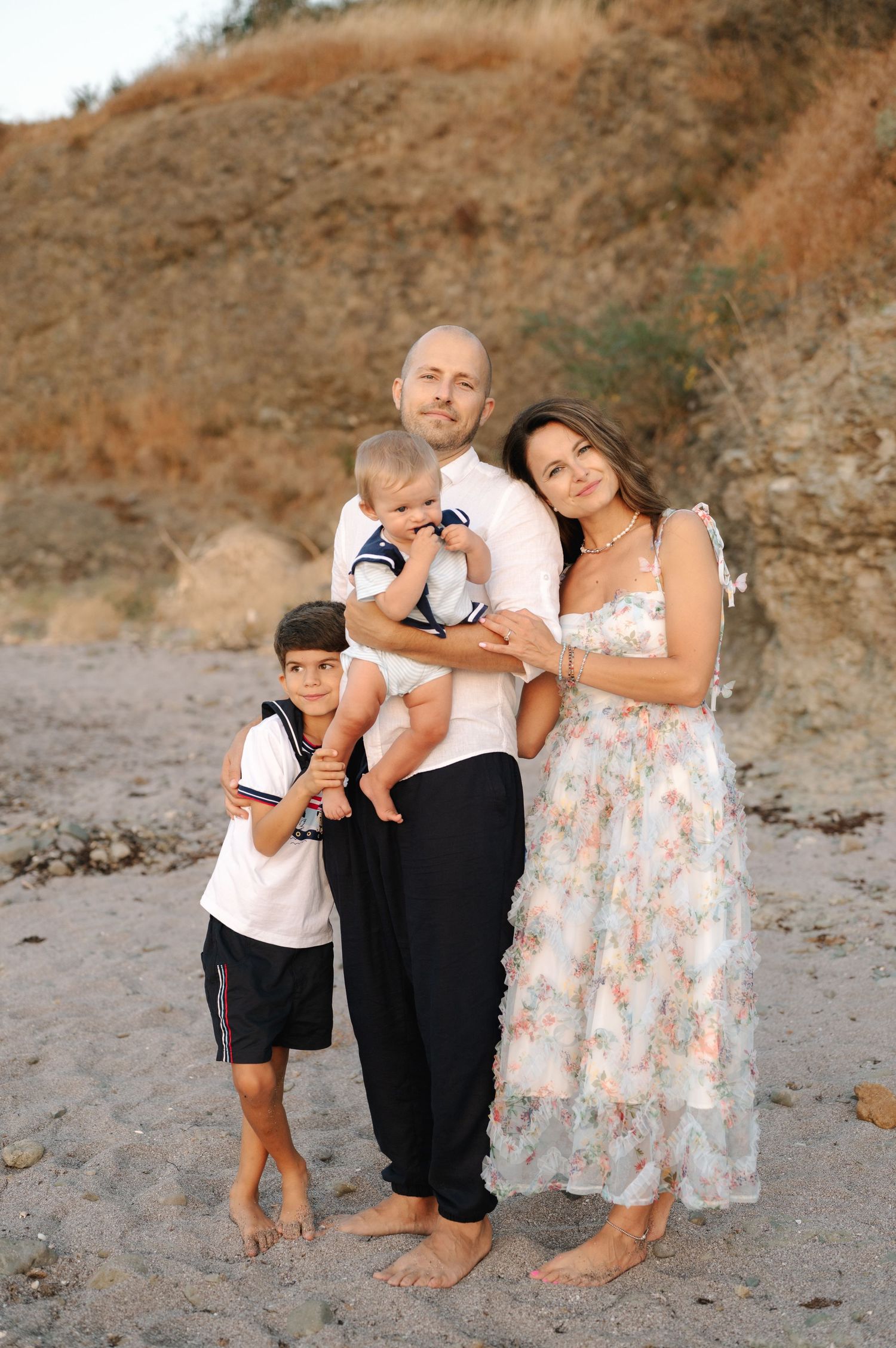 Family wearing white and floral outfits poses together on sandy beach at sunset in matching casual attire.