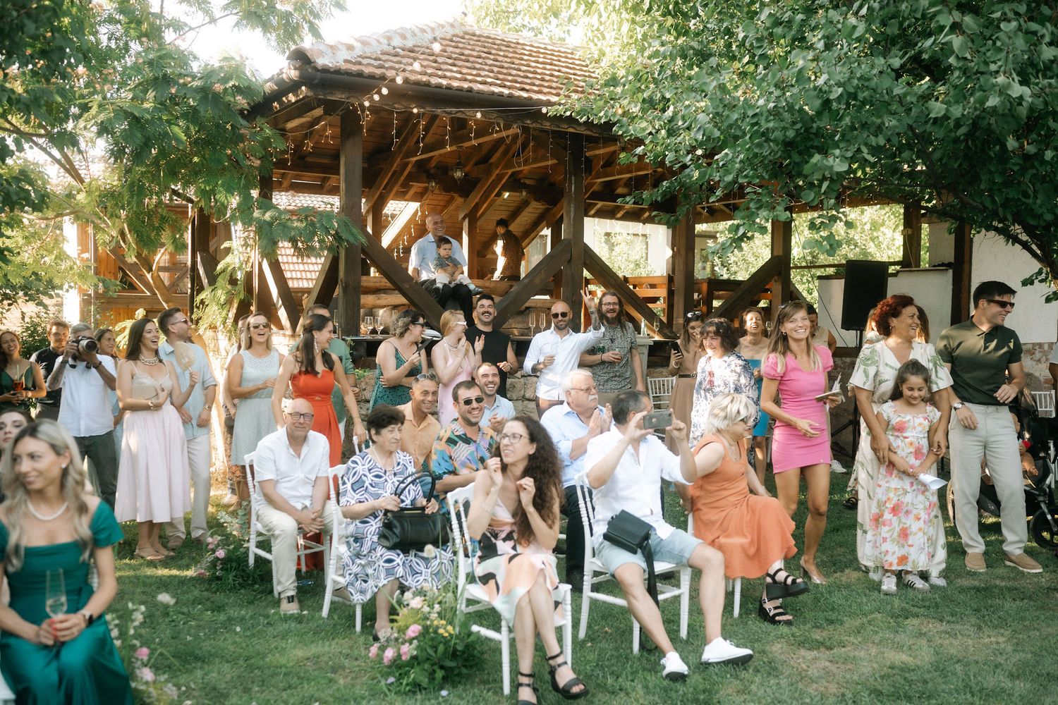 Outdoor wedding ceremony guests seated under a rustic wooden pavilion in a garden setting during summer.